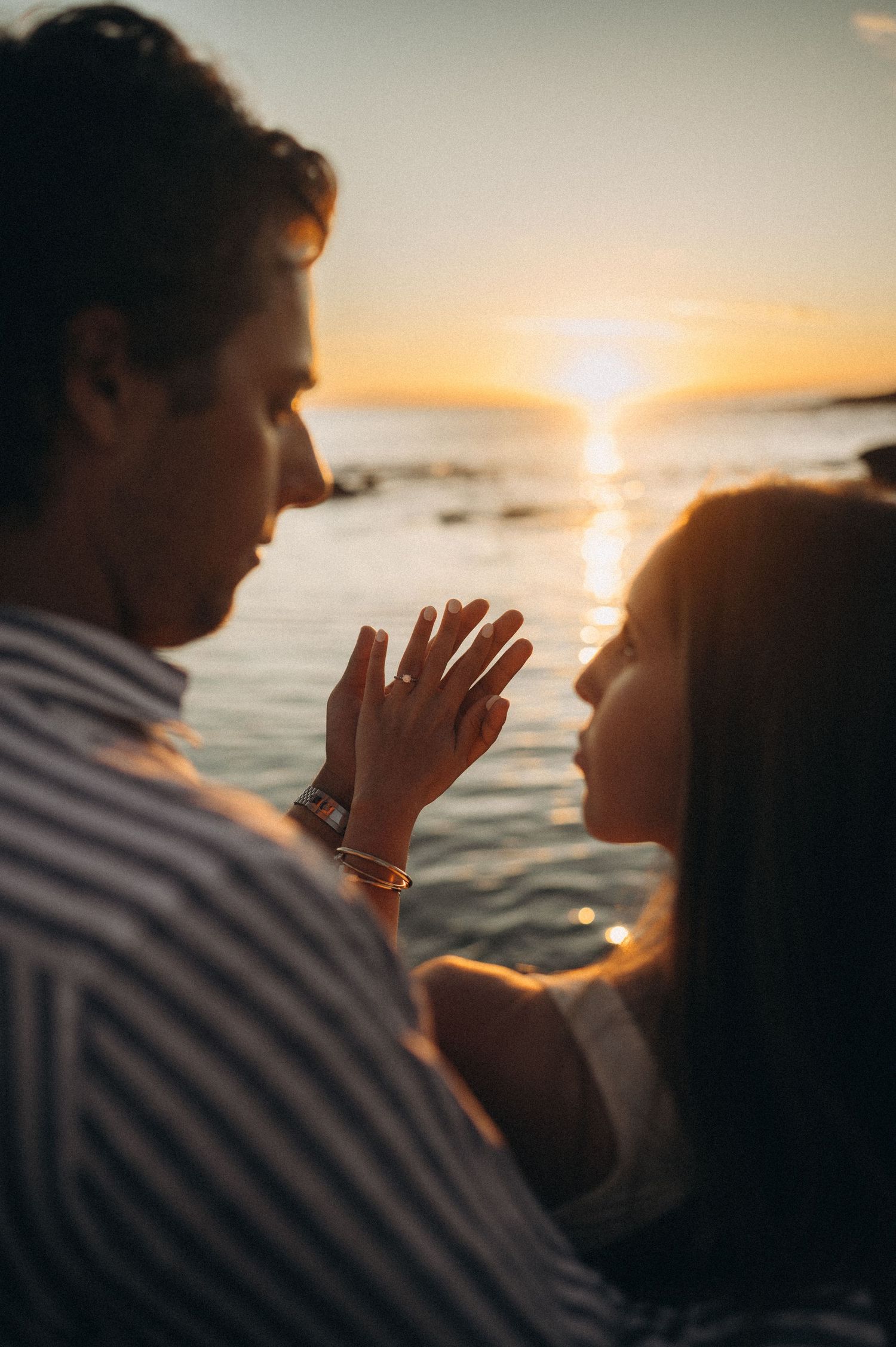 Couple sharing a romantic moment by the ocean at sunset, with woman showing off her engagement ring.