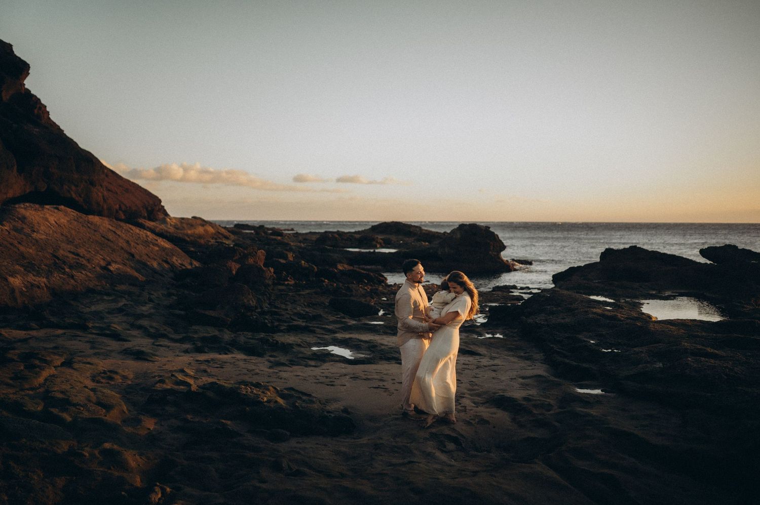 Two people in white clothing embrace on rocky volcanic coastline at sunset in Hawaii.