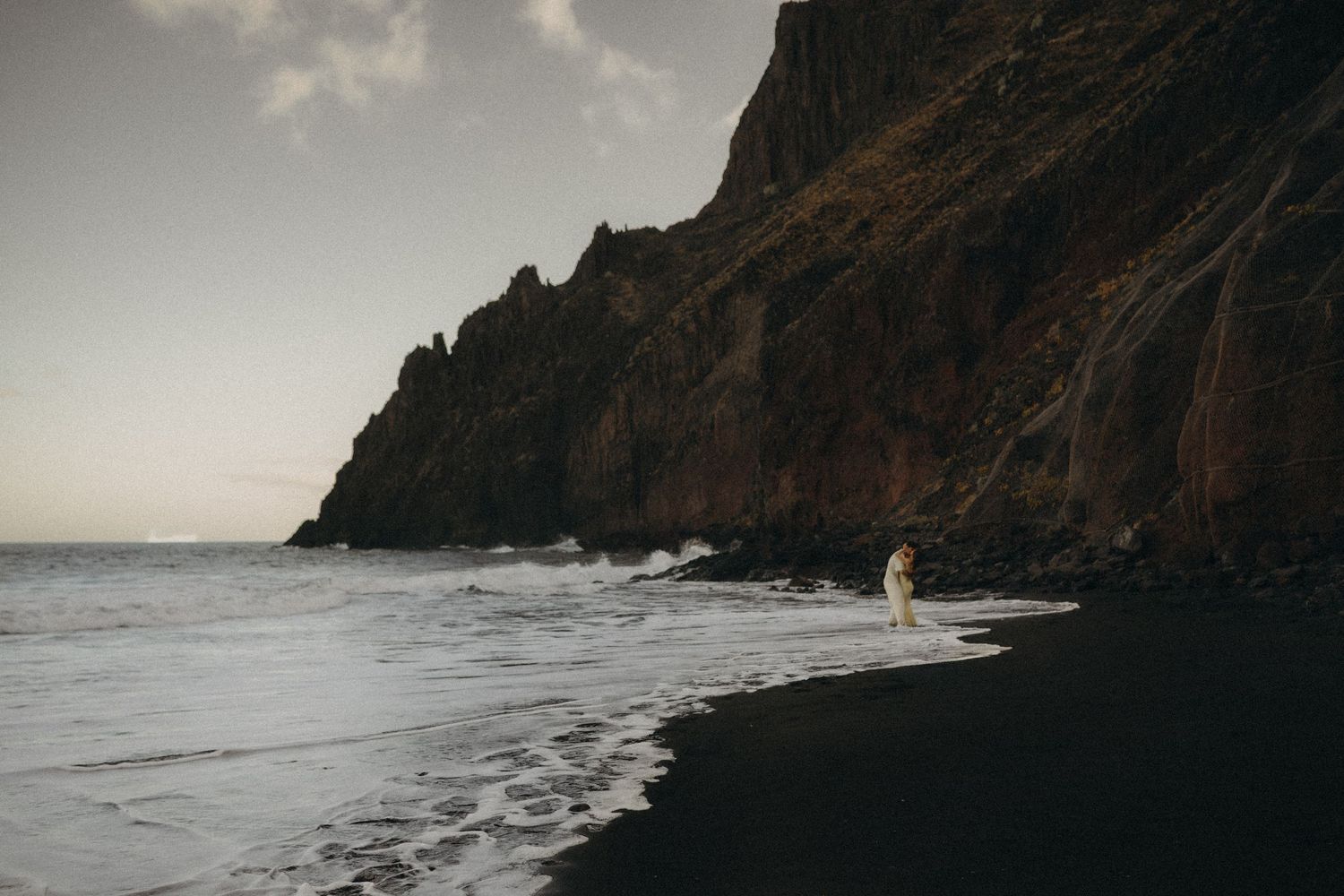 Person in white dress standing on black sand beach with dramatic rocky cliffs and ocean waves at dusk.
