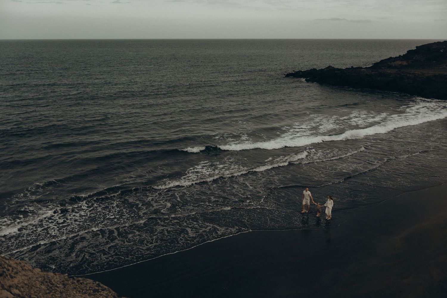 Two people walking on a dark volcanic beach with ocean waves and rocky coastline under overcast skies.