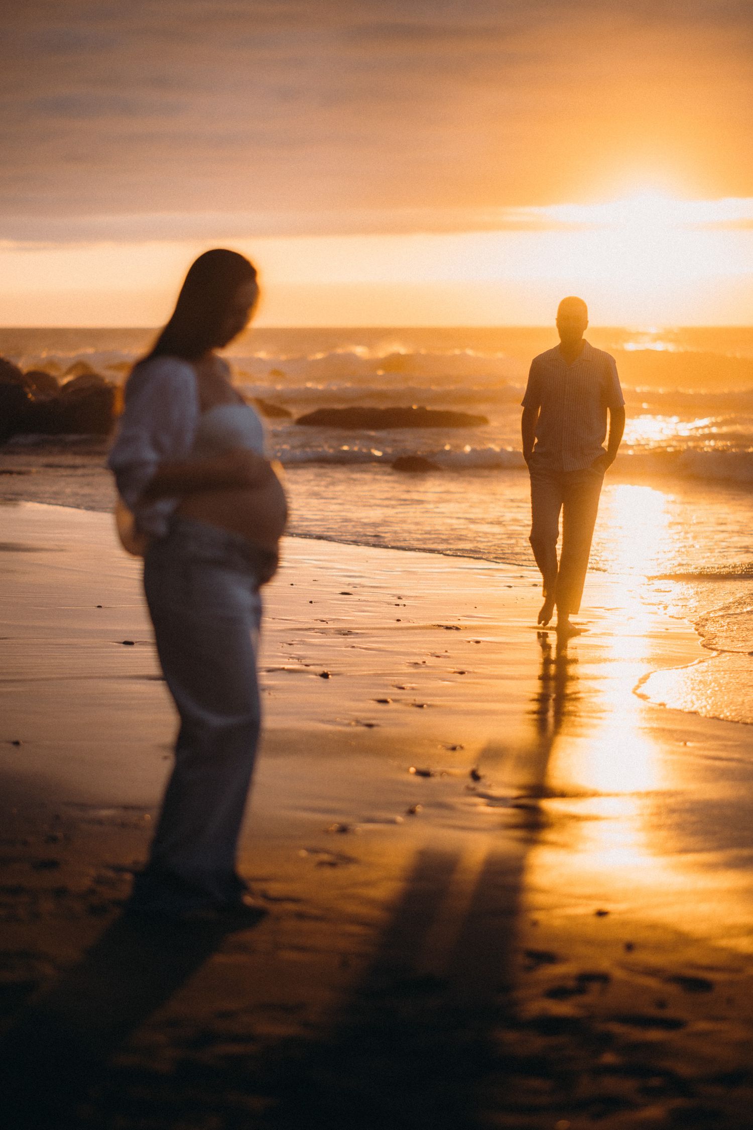 Pregnant woman cradling belly on beach at golden sunset while man walks toward ocean waves.