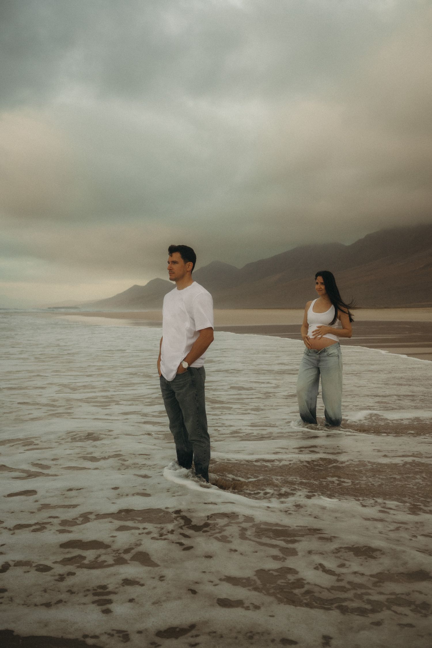 A couple stands in shallow ocean waves on a cloudy beach, with mountains in the background.