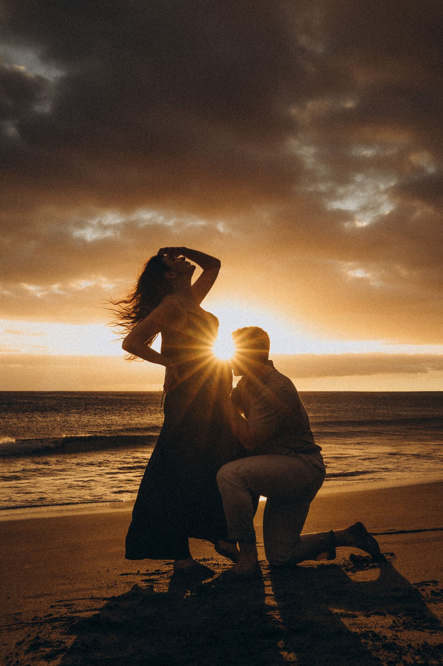 Romantic beach proposal at sunset with couple silhouetted against golden sky and ocean waves.