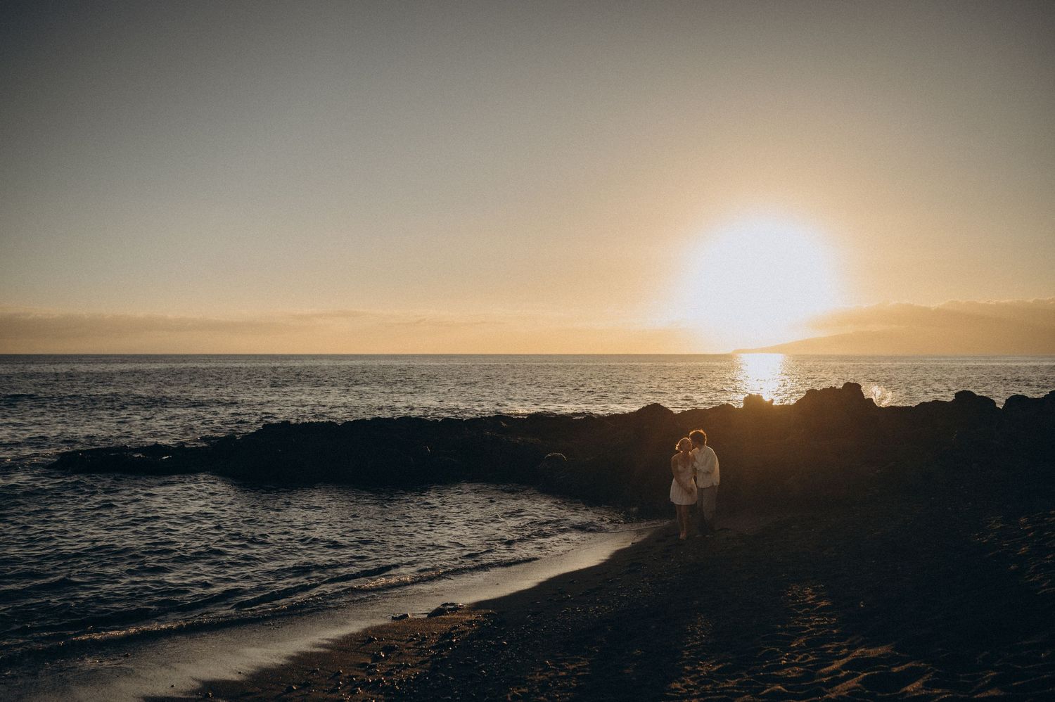 Couple embracing on rocky beach at golden sunset with ocean waves and glowing sun on the horizon.