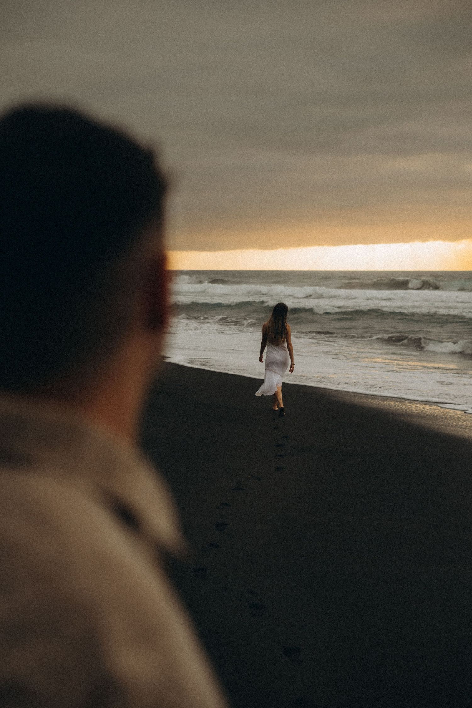 A woman in a white dress walks along a dark sandy beach at sunset while a man watches from the foreground.