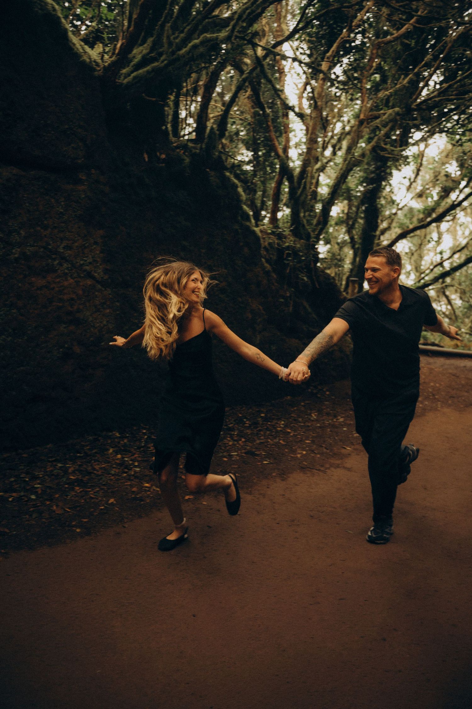 A joyful couple in matching black outfits holds hands while running along a forest path surrounded by ancient trees.