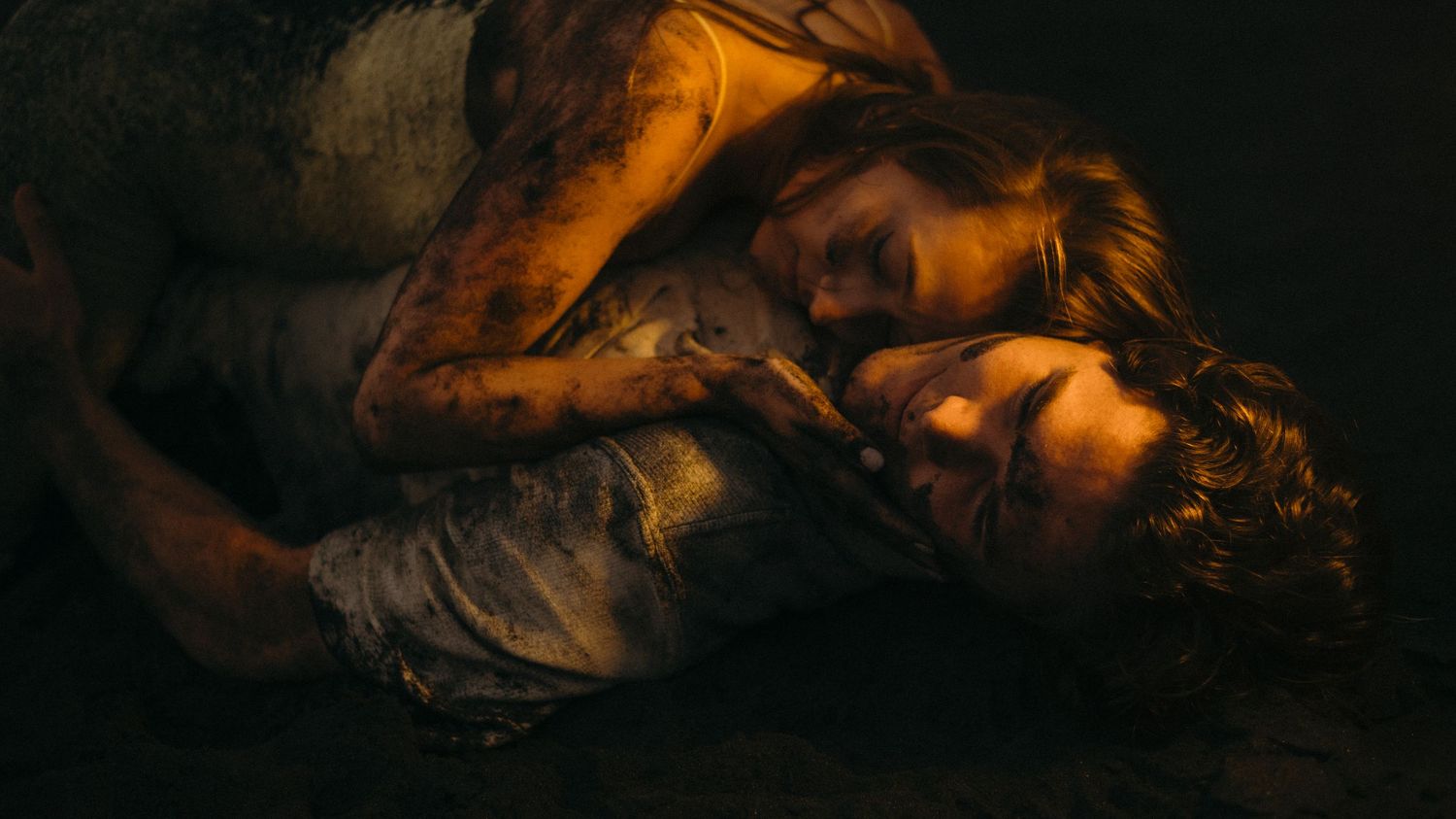 Two mud-covered figures lying together in a dark, dramatic scene with cinematic lighting.