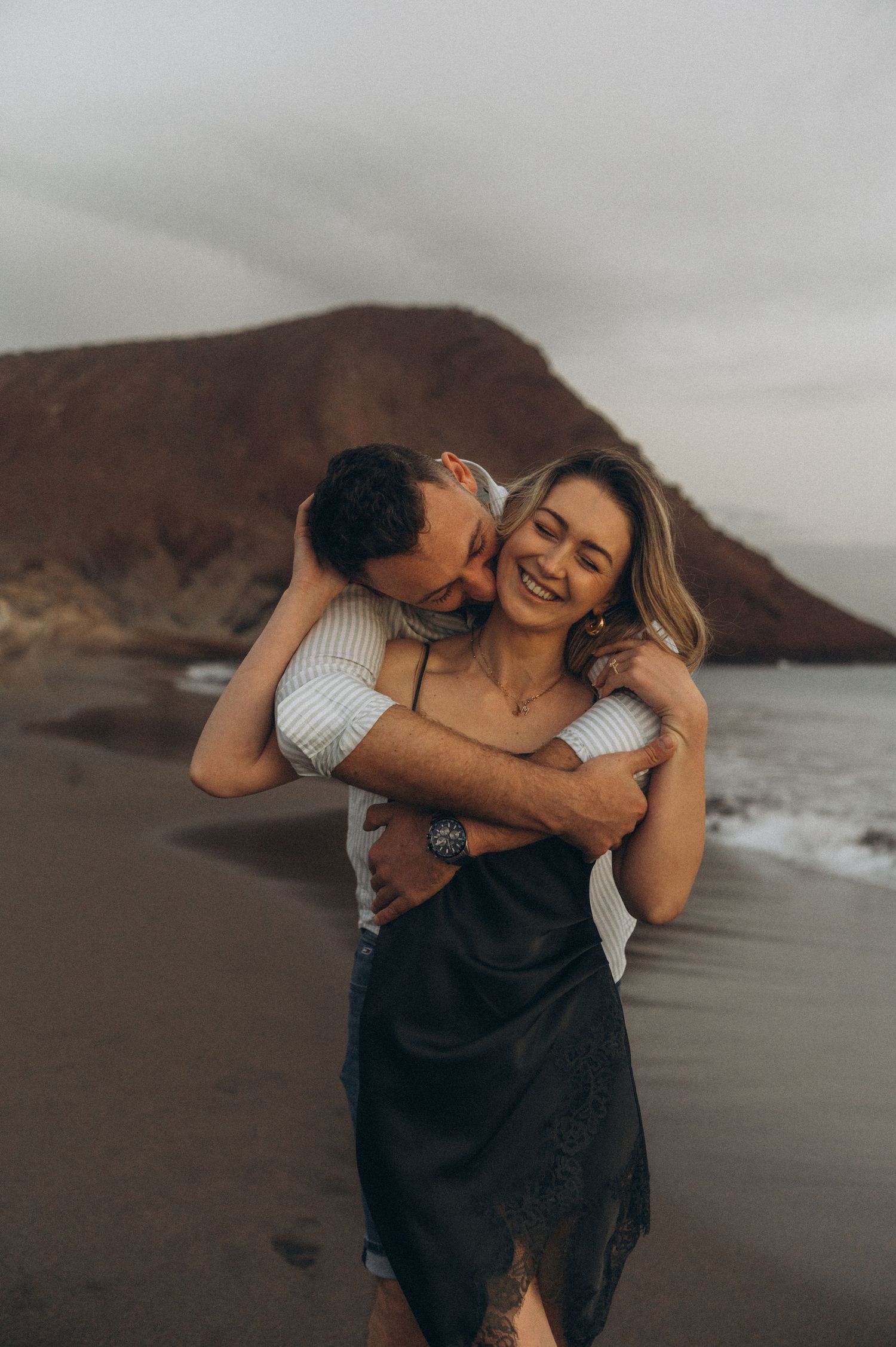 Man holding and kissing his partner in embrace with Montaña Roja in background, Playa de La Tejita, Tenerife