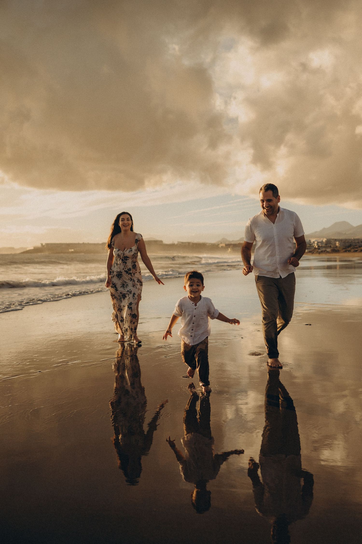 A family enjoys a sunset stroll along a reflective beach with dramatic cloudy skies in the background.