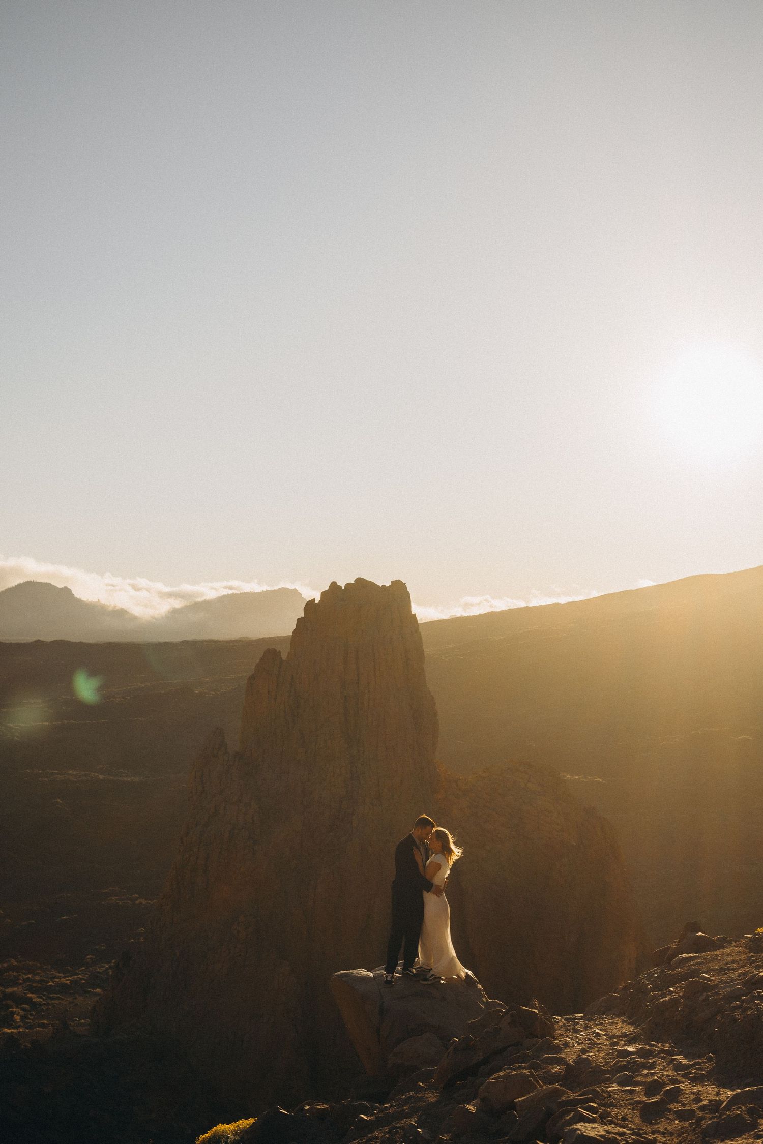 Bride and groom embrace on rocky mountain summit at golden sunset, dramatic landscape backdrop.