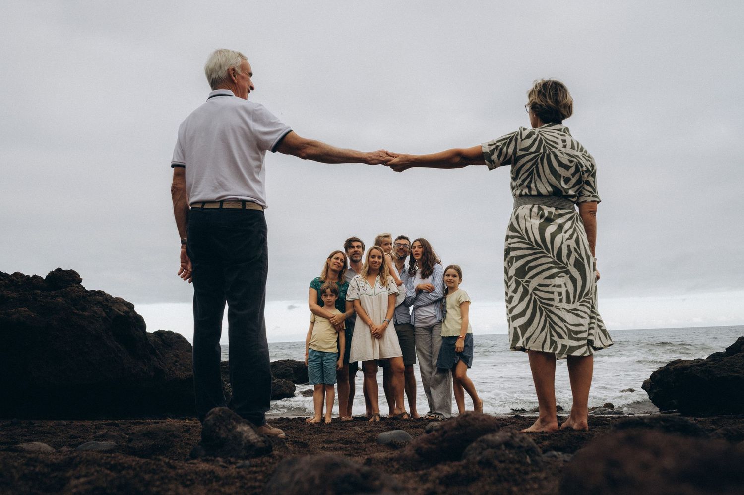 Grandparents holding out hands creating a frame with family in foreground on black sand at Los Refugios