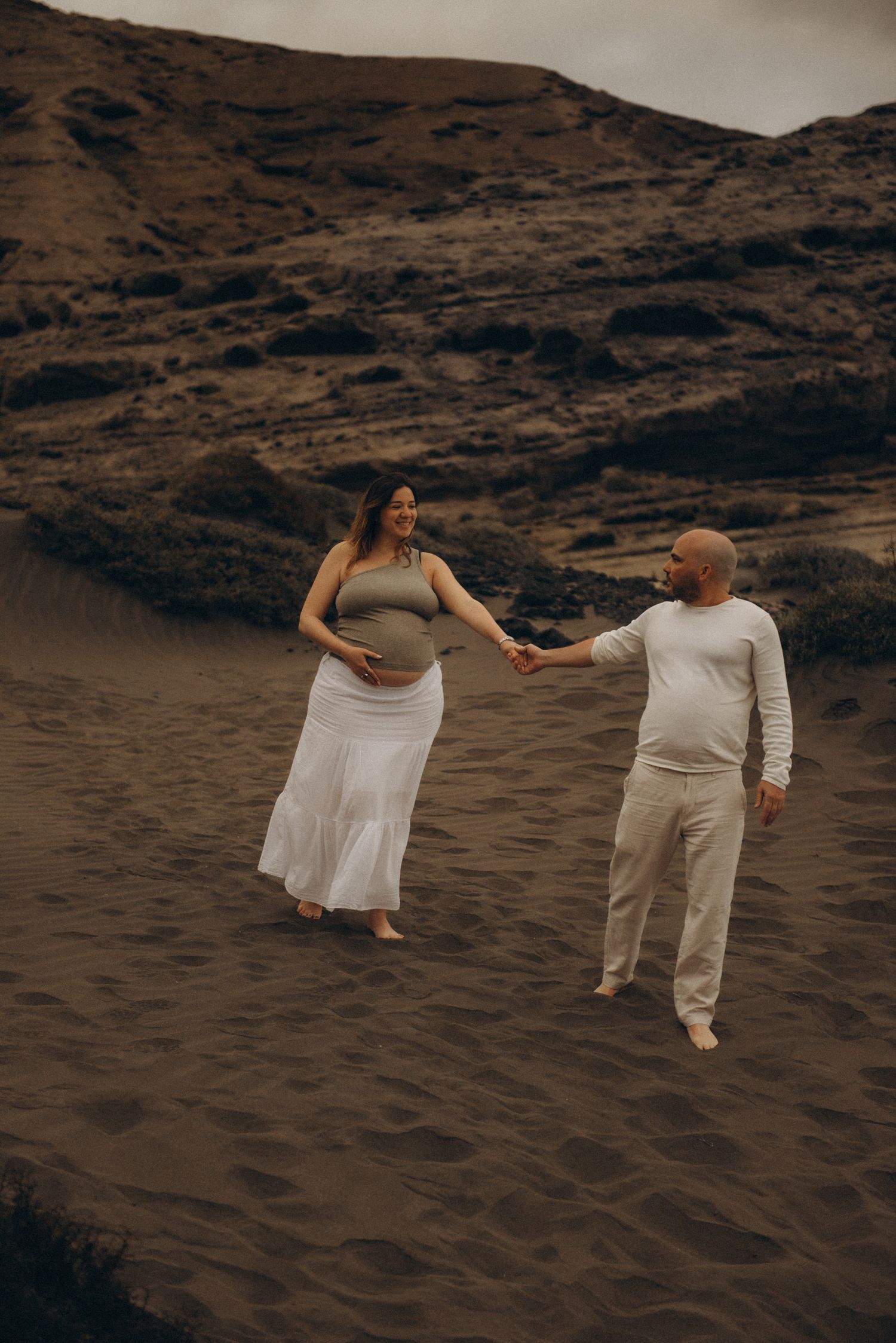 Pregnant couple holding hands and walking on sandy dunes, woman in white skirt and crop top, man in beige outfit.