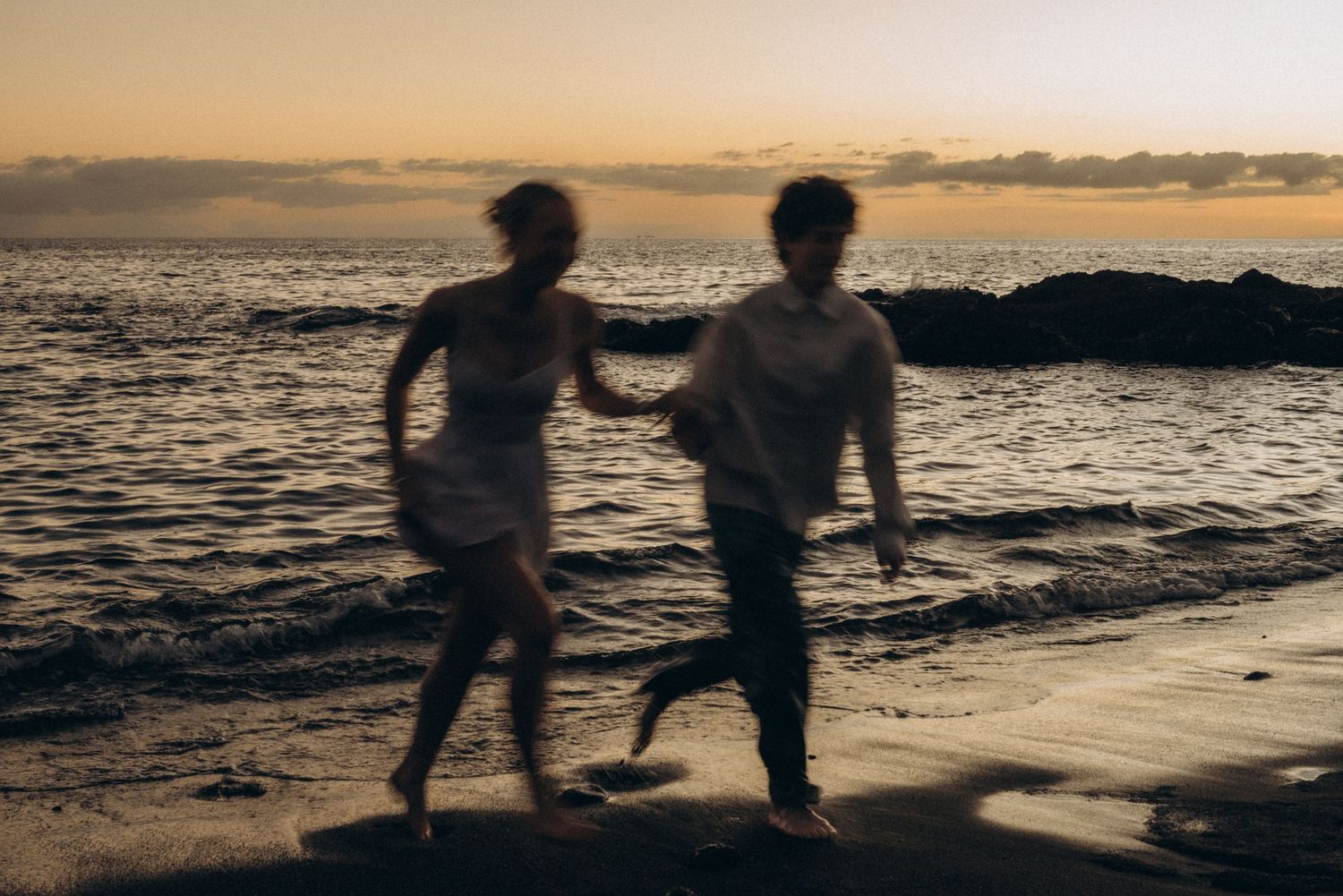 Blurred couple running on a sandy beach at sunset with ocean waves and dramatic sky in background.