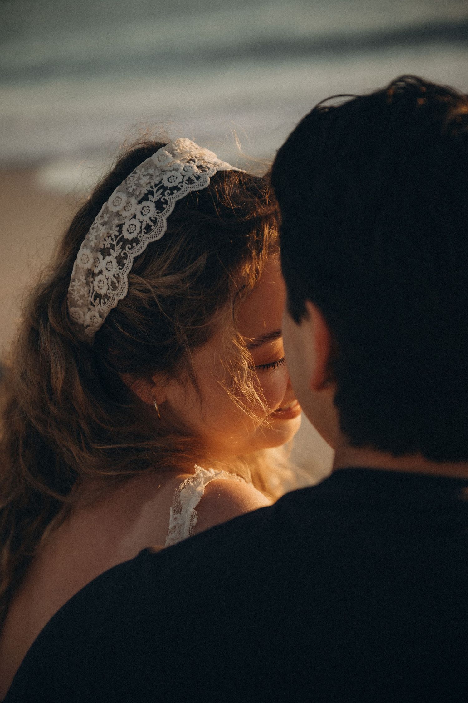 A bride wearing a lace headband smiles softly near the ocean at golden hour with her partner.