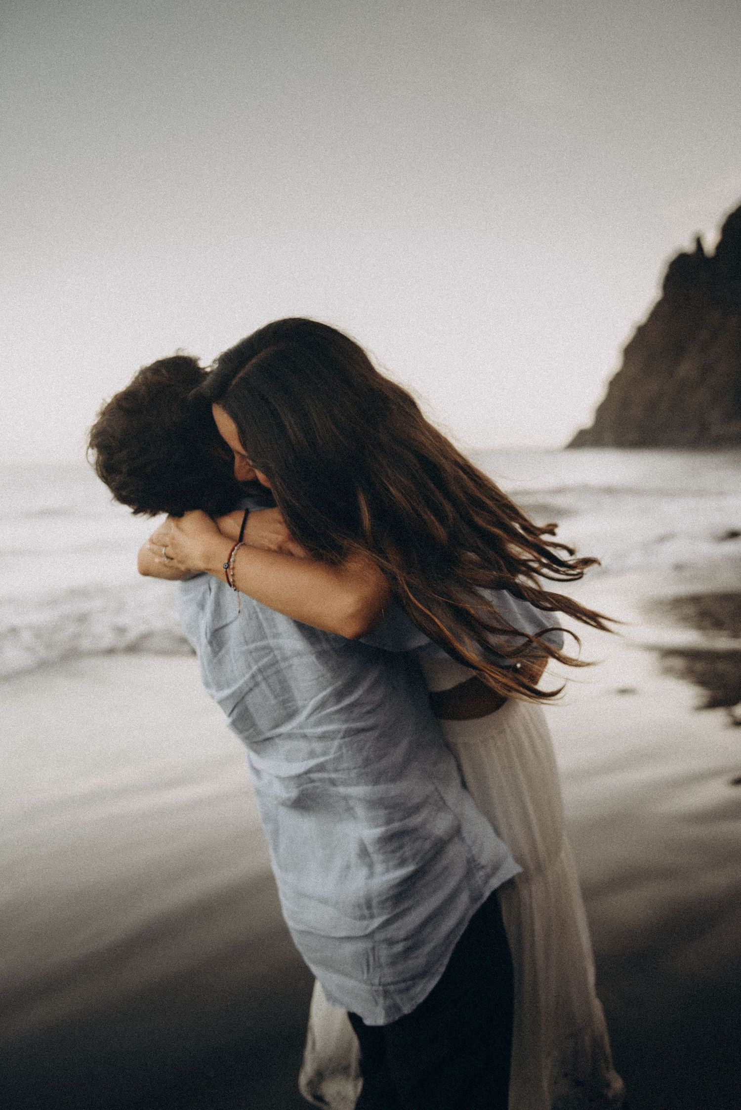Couple embracing romantically on a misty beach, woman with long hair lifted by man in blue shirt near ocean waves.