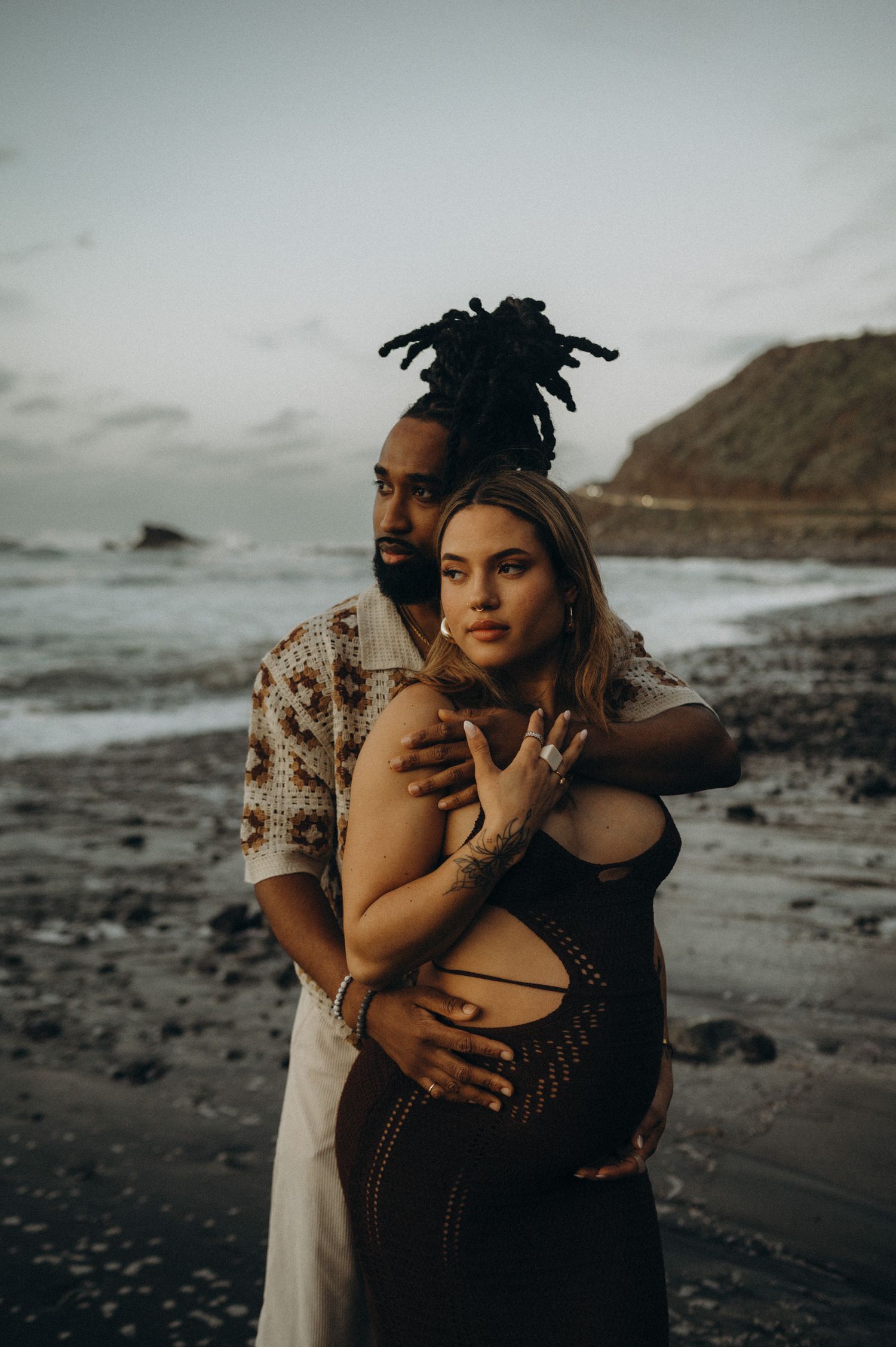 Couple sharing a tender embrace on a rocky beach at dusk, with ocean waves and cliffs in the background.