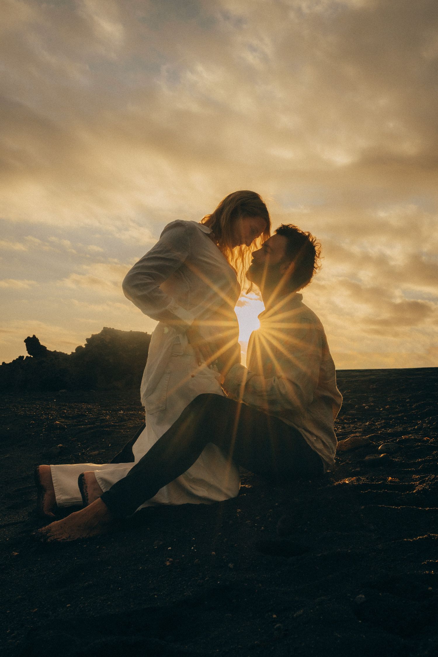 Couple sharing intimate moment silhouetted against stunning sunset with golden sun flare on dark beach.