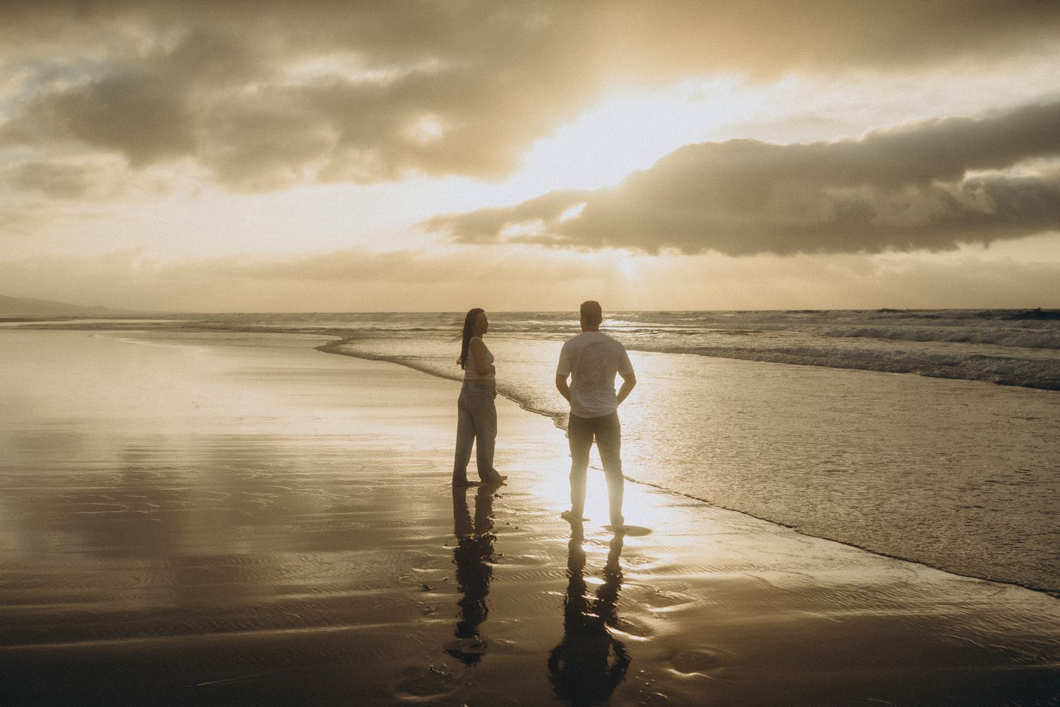 Couple holding hands on a beach at golden sunset with dramatic cloudy sky and ocean waves reflecting warm light.
