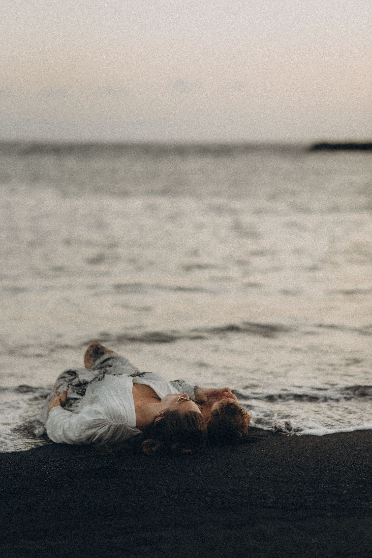 Mother lying in embrace by ocean with toddler beside her on black sand, Playa del Duque