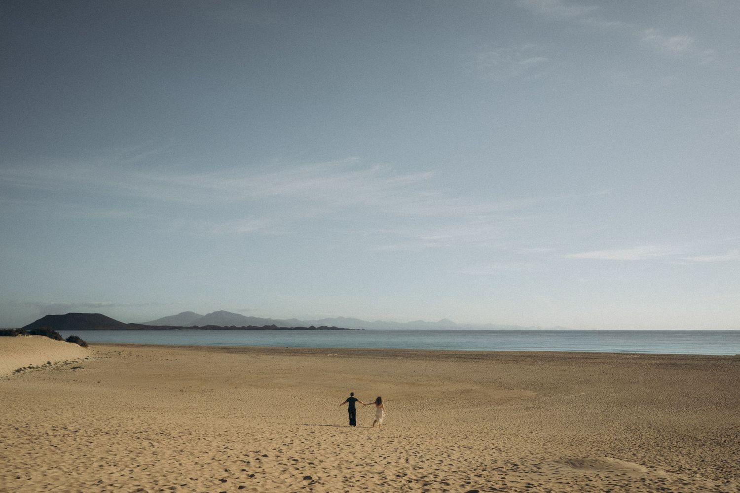 Two people stand on a vast sandy beach with calm turquoise waters and distant mountains under a clear blue sky.