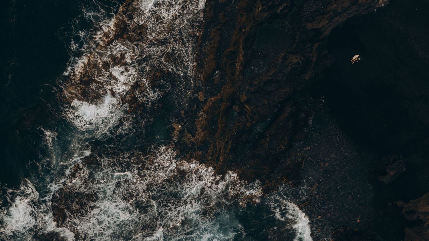 Aerial view of dark ocean waves crashing against rocky coastal cliffs, with white sea foam and a small boat visible.