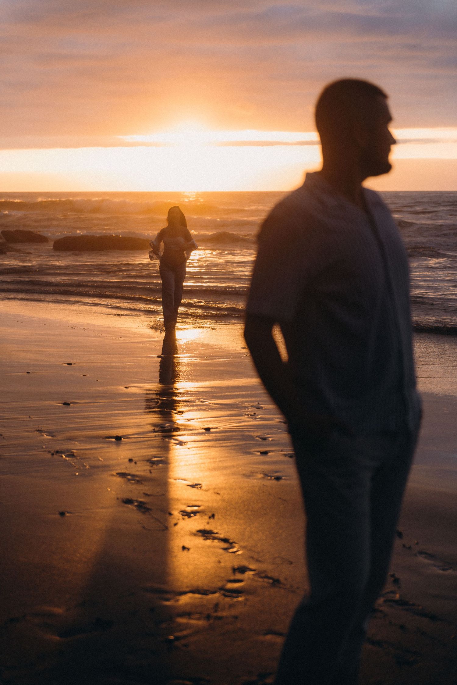 Silhouette of a man on a beach at golden sunset with a woman walking toward glowing ocean waves.
