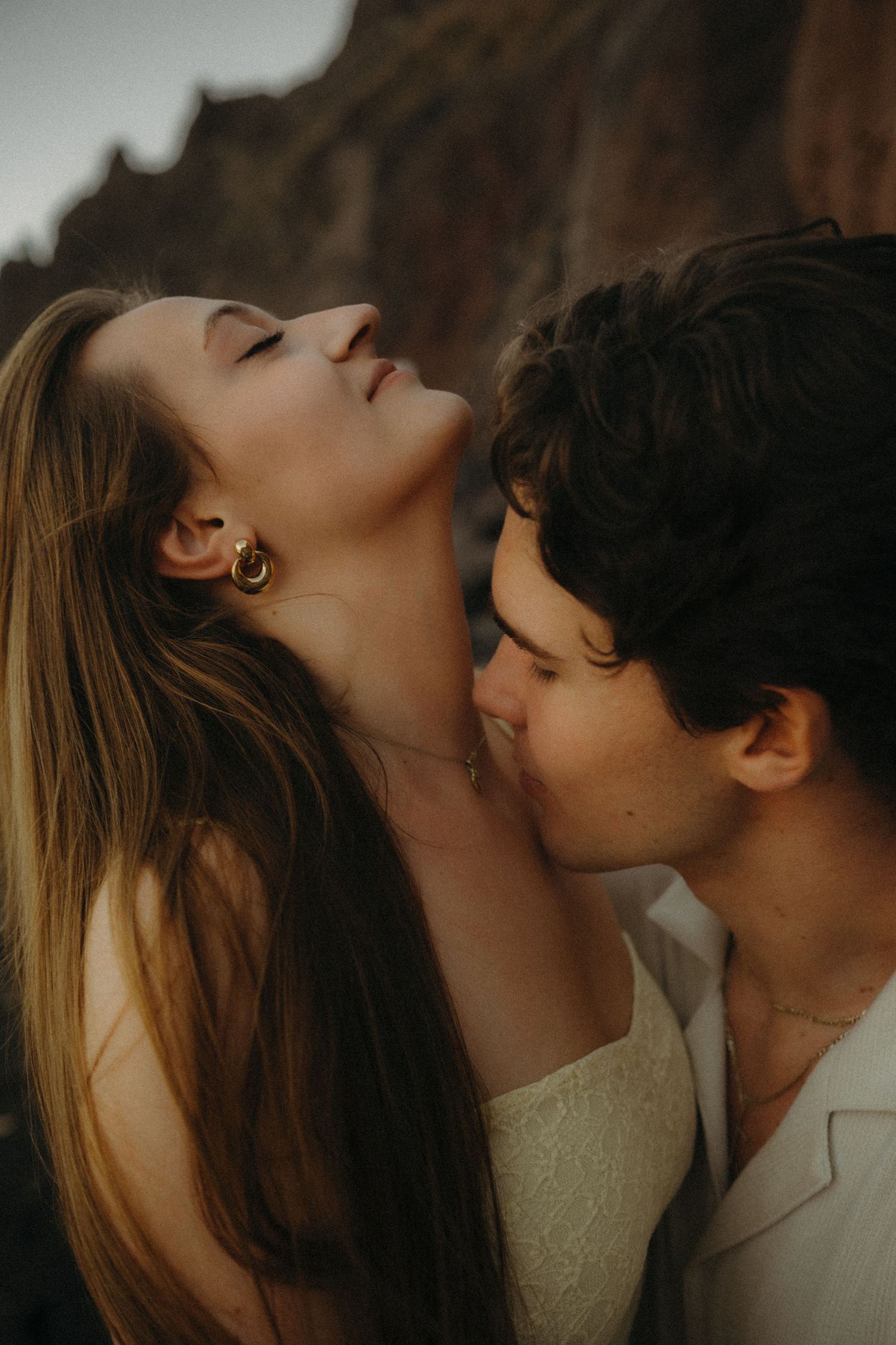 A couple shares an intimate romantic moment outdoors, with the man kissing the womans neck as she tilts her head back.