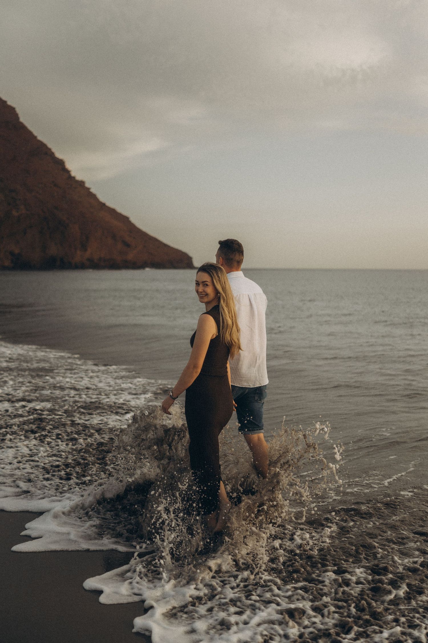 A couple splashes through waves along a beach with Montana Roja at Playa de La Tejita, Tenerife