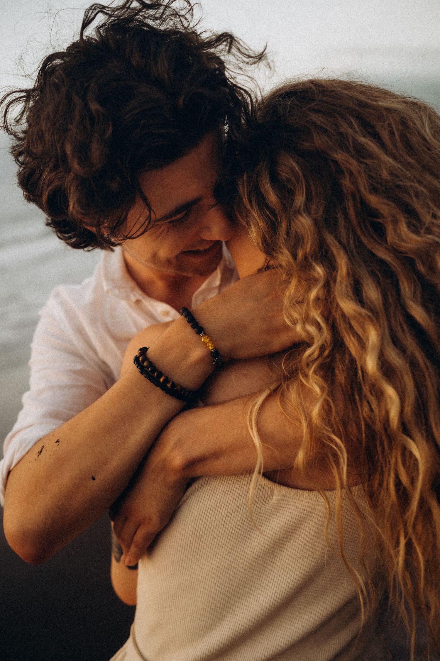 A couple with curly hair shares a romantic embrace near the water, the man wearing a black beaded bracelet.