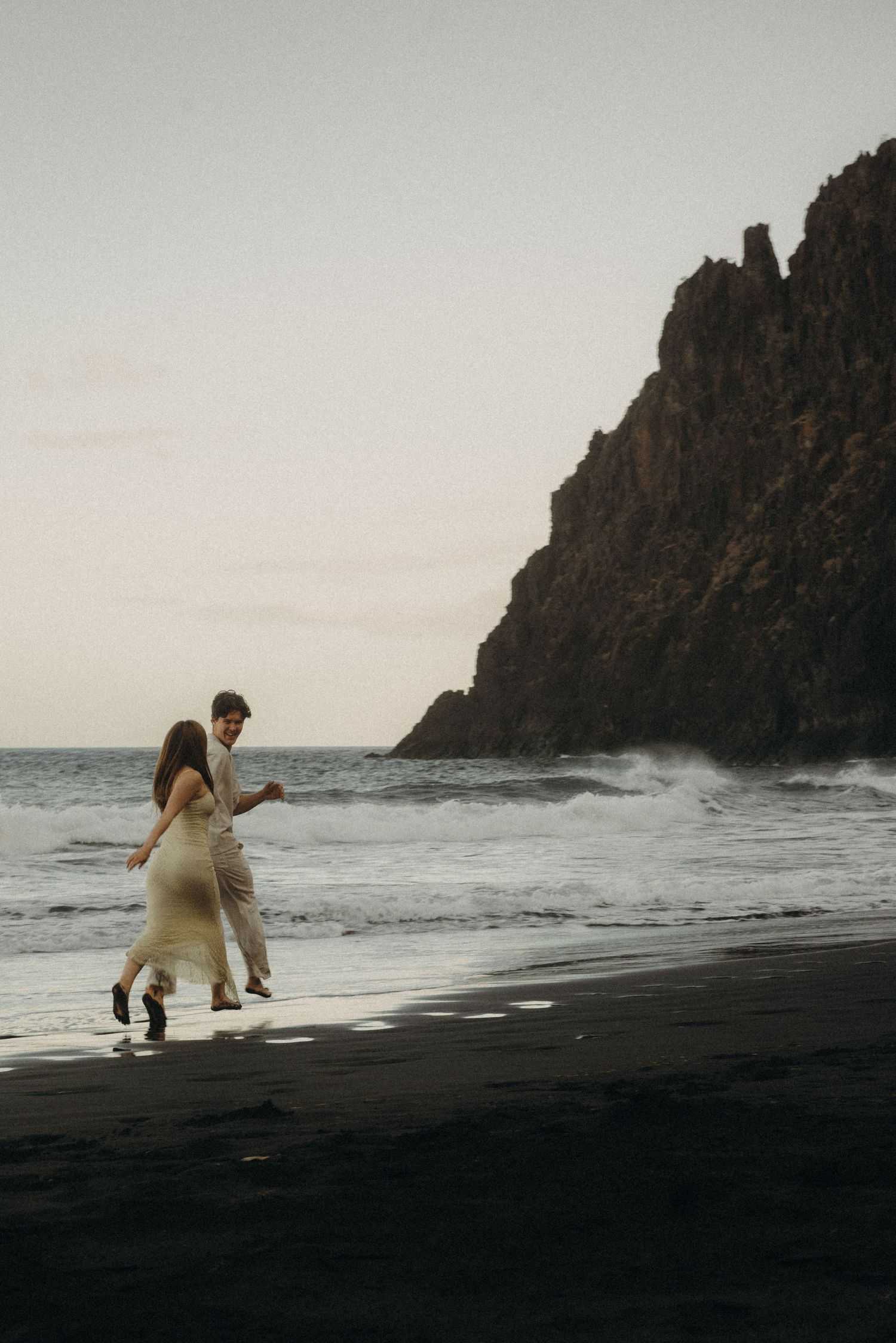Couple running along a black sand beach at dusk with dramatic rocky cliffs and crashing ocean waves.