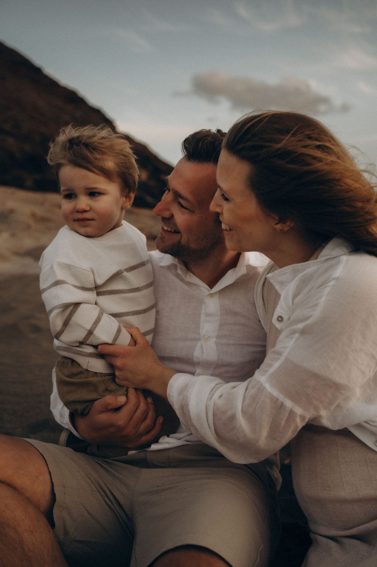 A happy family of three sits on a beach, parents smiling at their toddler in a striped sweater. Photoshoot at Playa de La Tejita, Tenerife