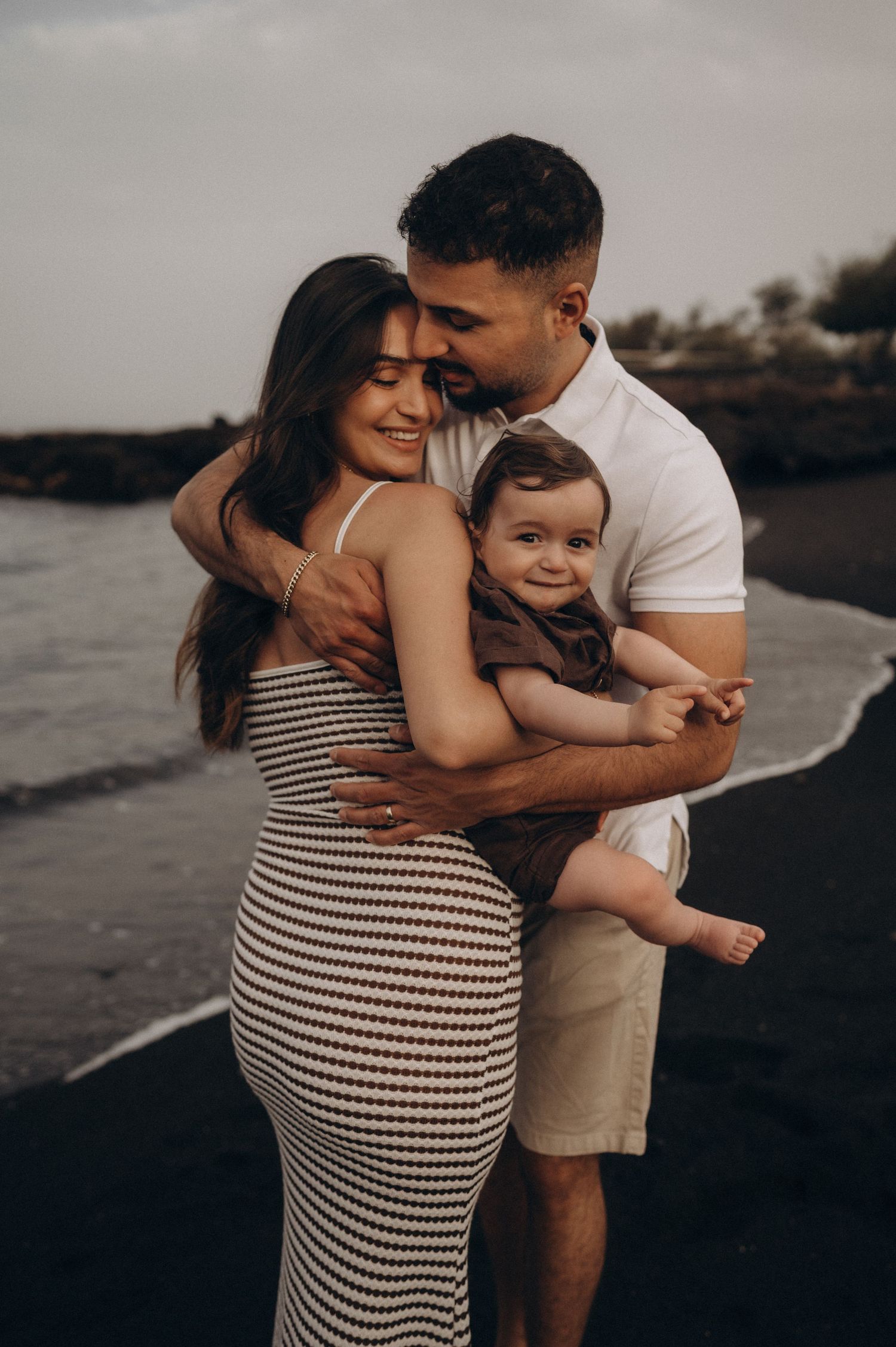 Happy young family with baby on black sand beach, parents embracing and smiling during golden hour photo session.