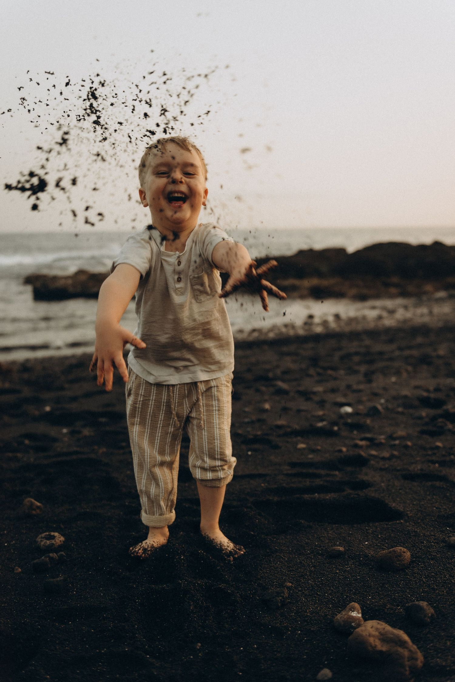 Joyful toddler laughing and throwing dark sand on a rocky beach at dusk, covered in dirt.