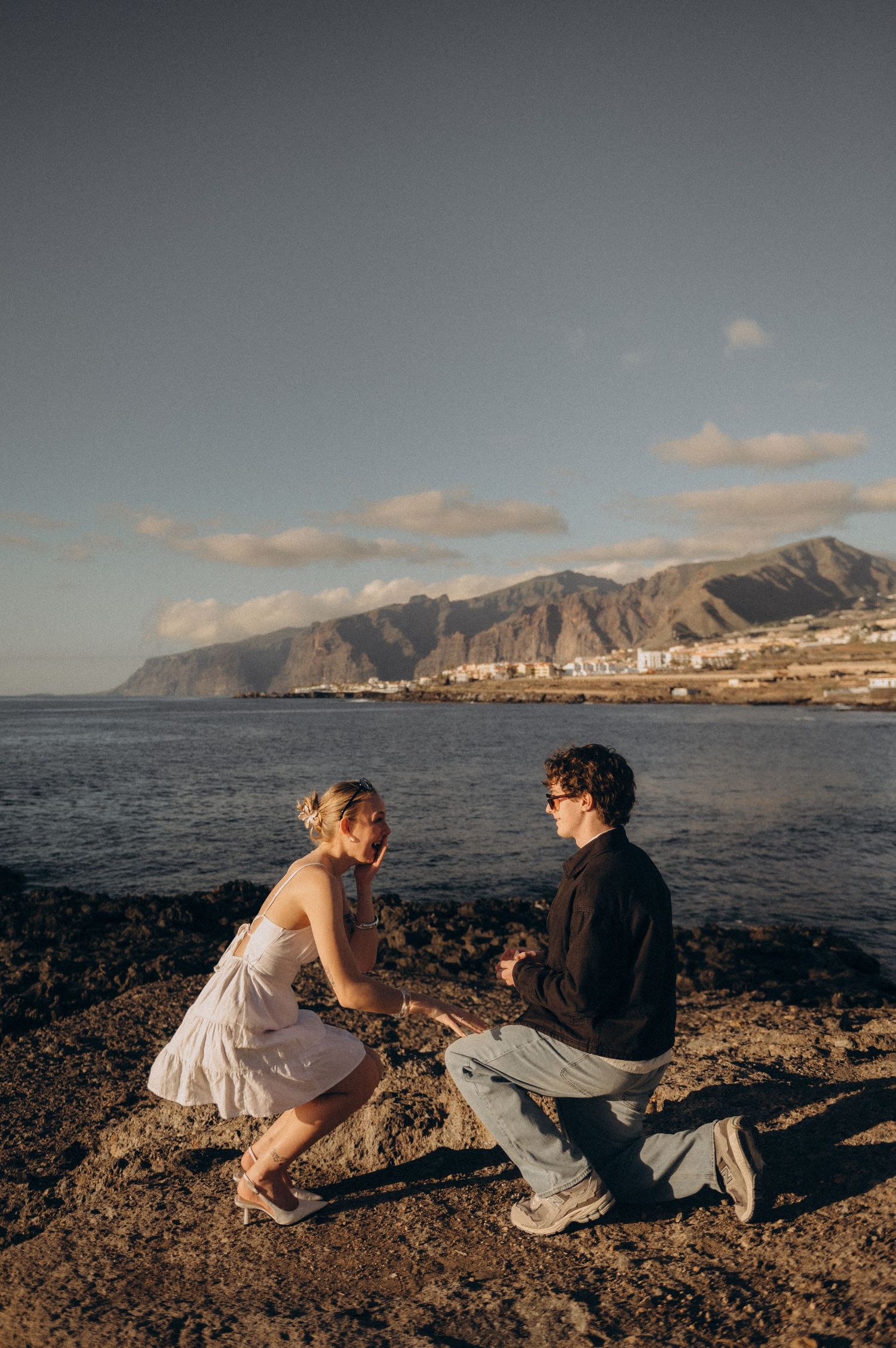 Man proposing to woman on rocky coastline with mountains and ocean in background at golden hour.