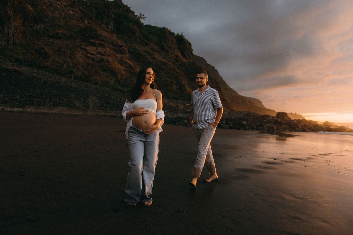 Pregnant couple walking on black sand beach at sunset with dramatic cliffs and golden sky backdrop.