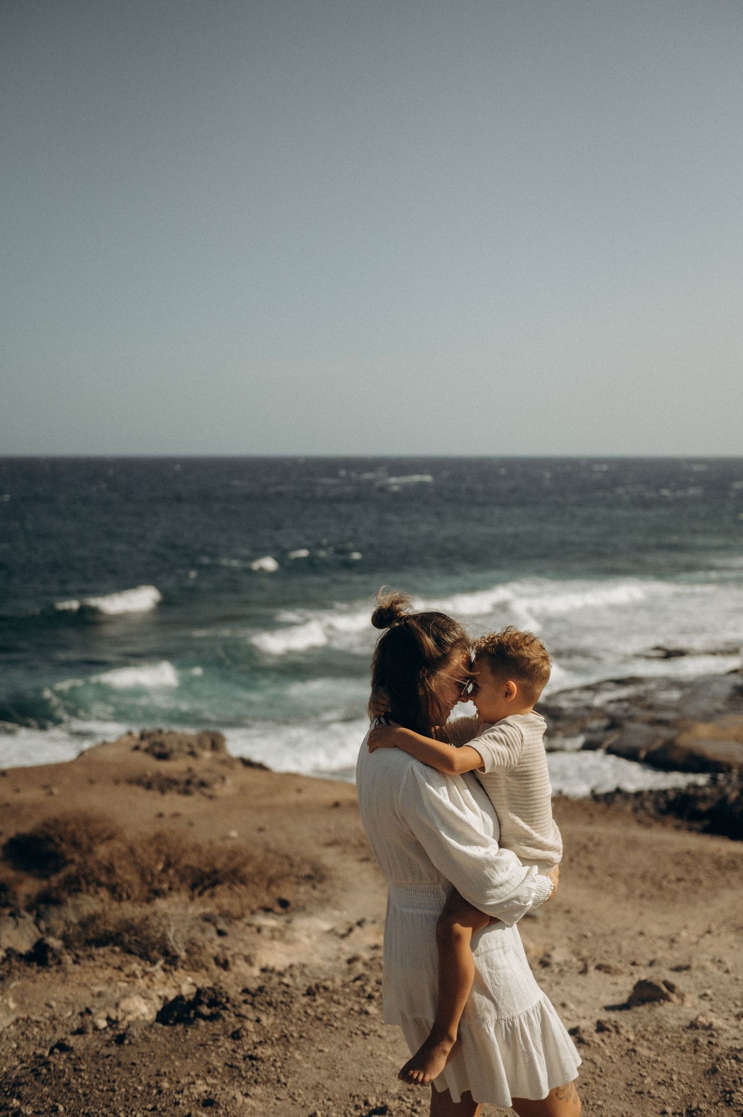 Mom and son embracing on coastal cliff overlooking ocean waves on moody overcast day during Tenerife family photoshoot.