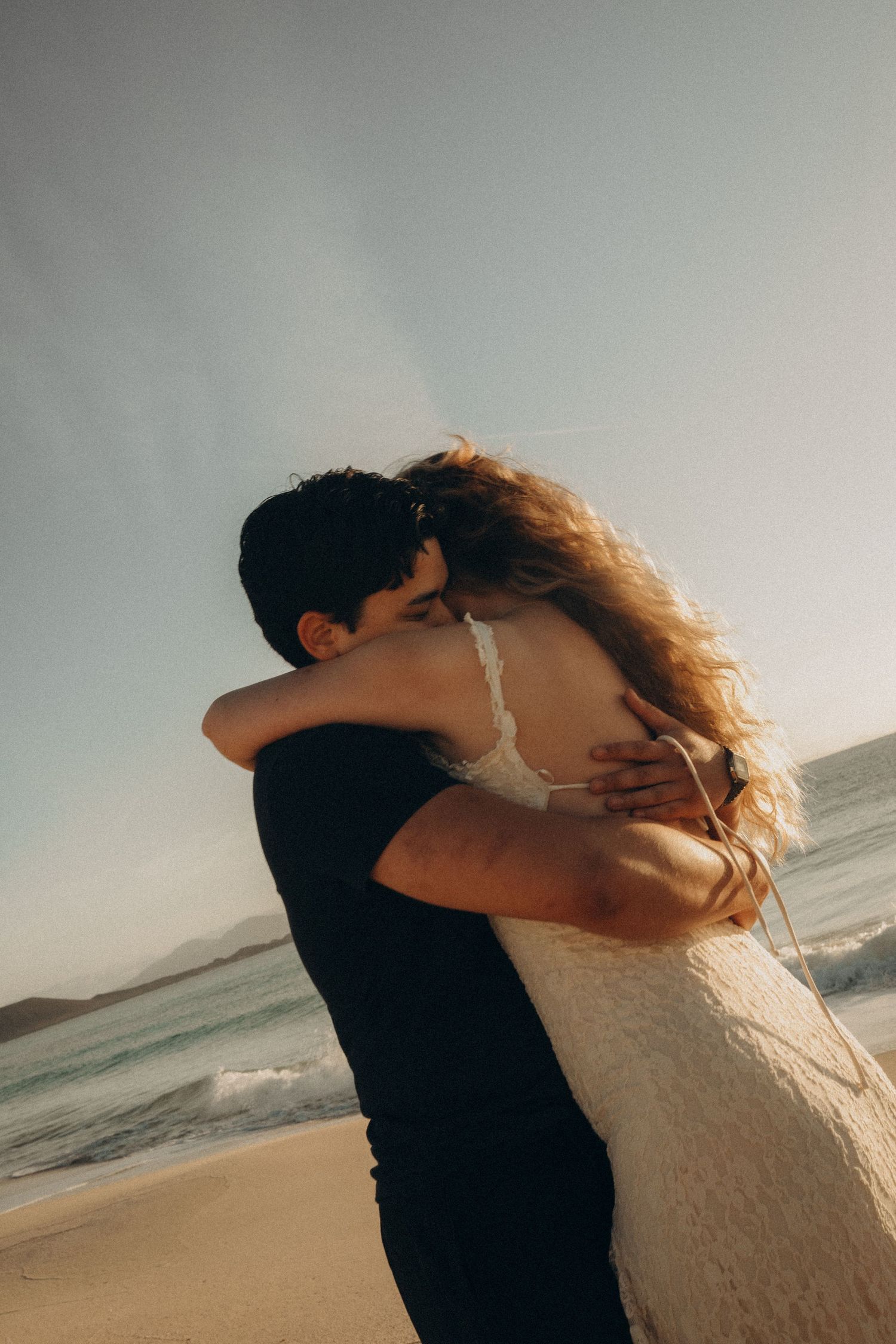 Couple embracing romantically on a sandy beach with ocean waves and mountains in the background on a sunny day.
