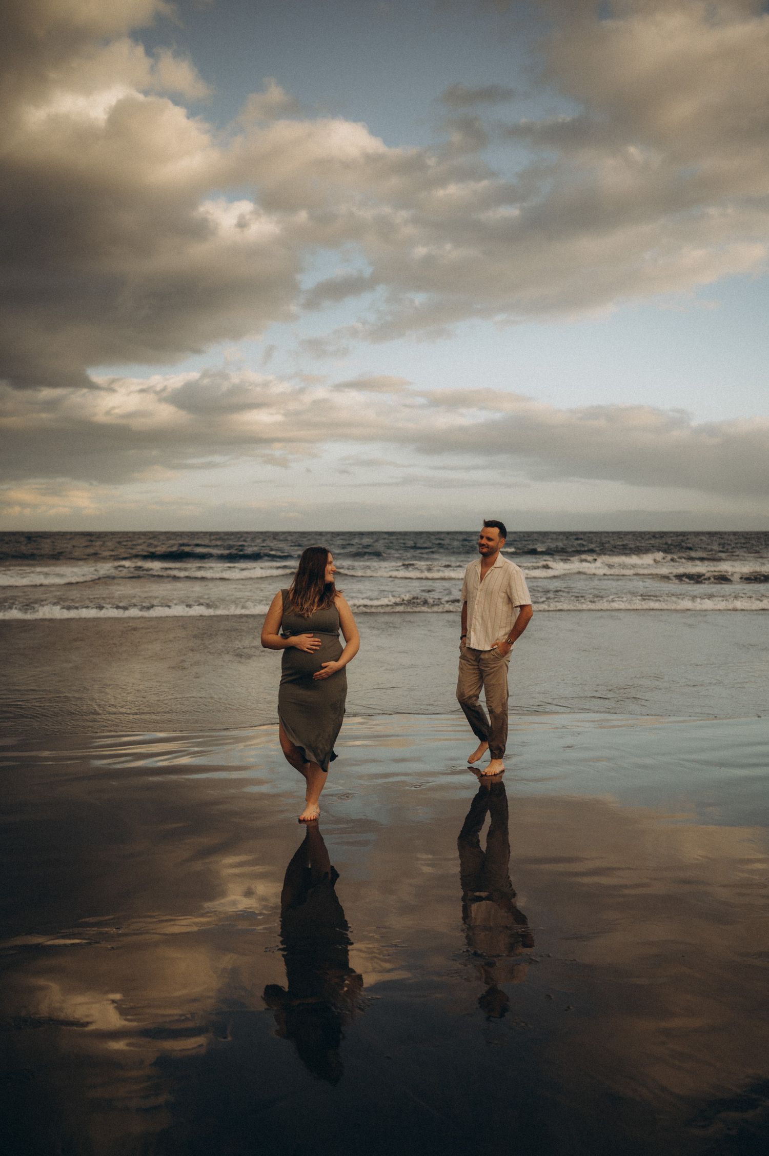 Pregnant couple walking on reflective wet beach at sunset with dramatic cloudy sky and ocean waves behind them.