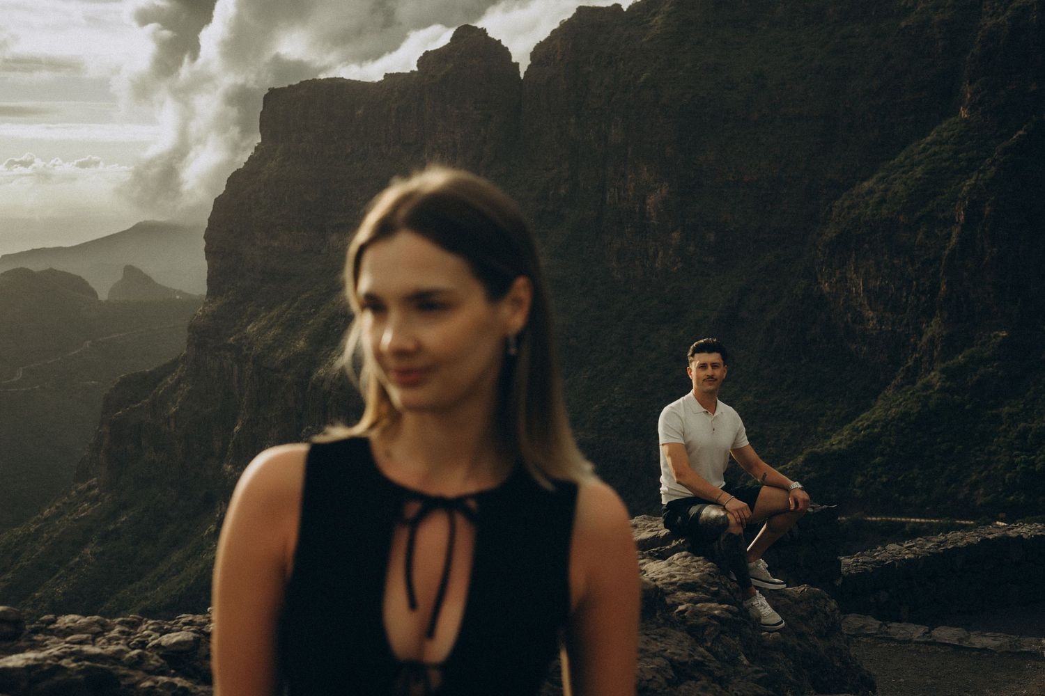 Couple posing on rocky mountain cliff with dramatic cloudy sky, woman in black dress foreground, man in white shirt background.