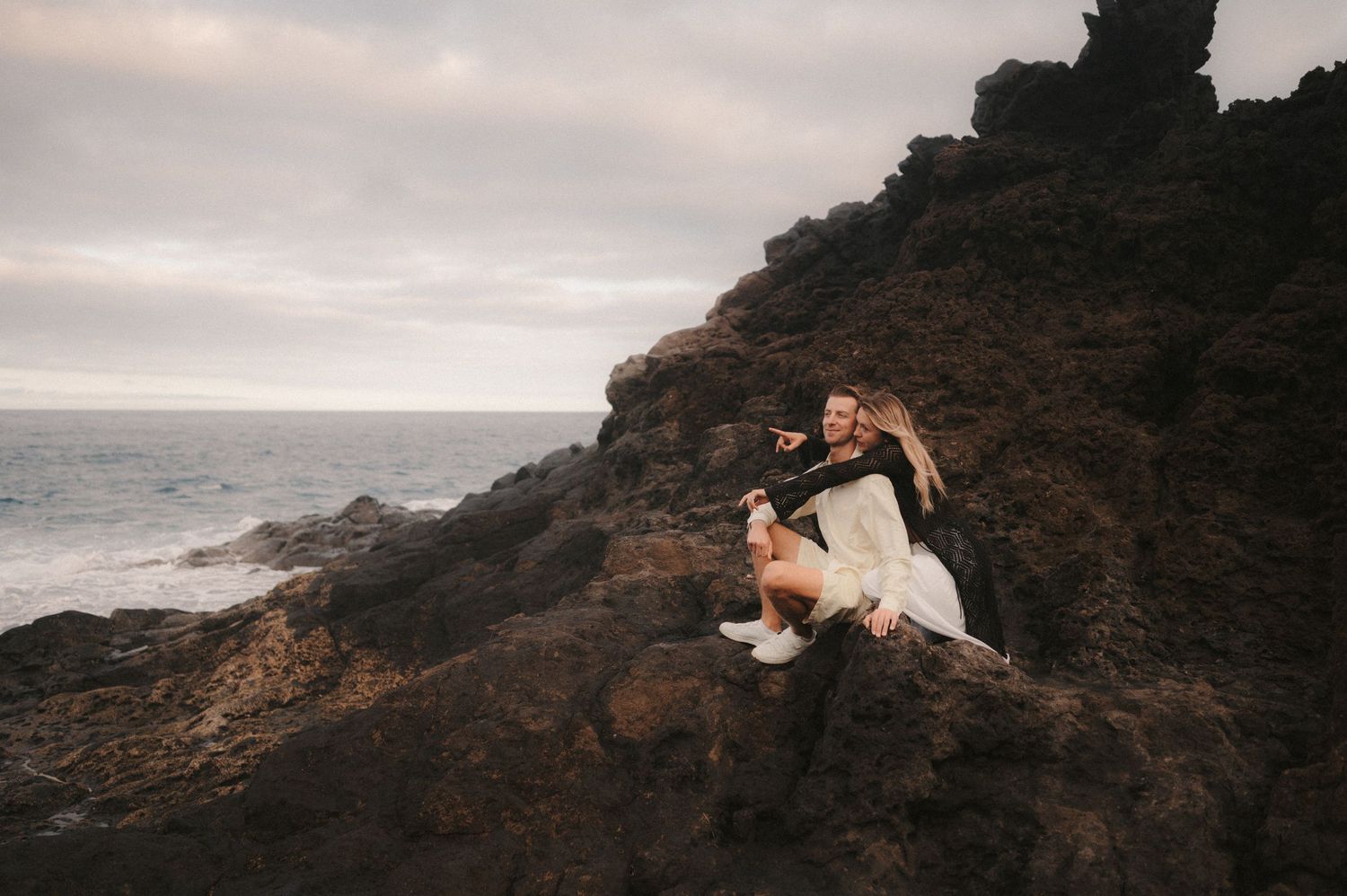 Young couple sitting on rocks by the ocean, enjoying the ocean view under Montaña Roja, Tenerife