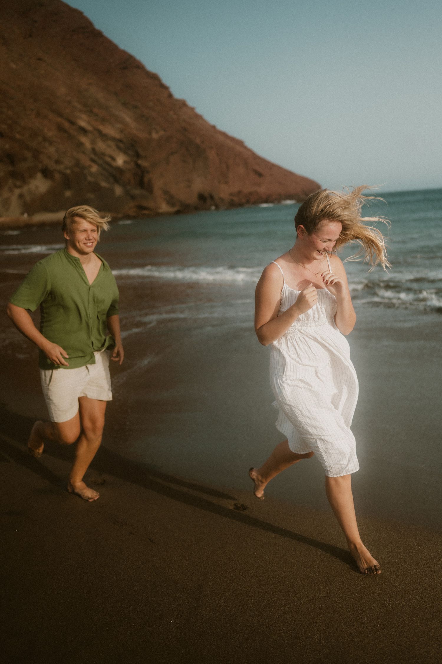 Young couple running hand in hand barefoot on Playa Tejita beach beside the ocean during sunset, Tenerife couples photoshoot