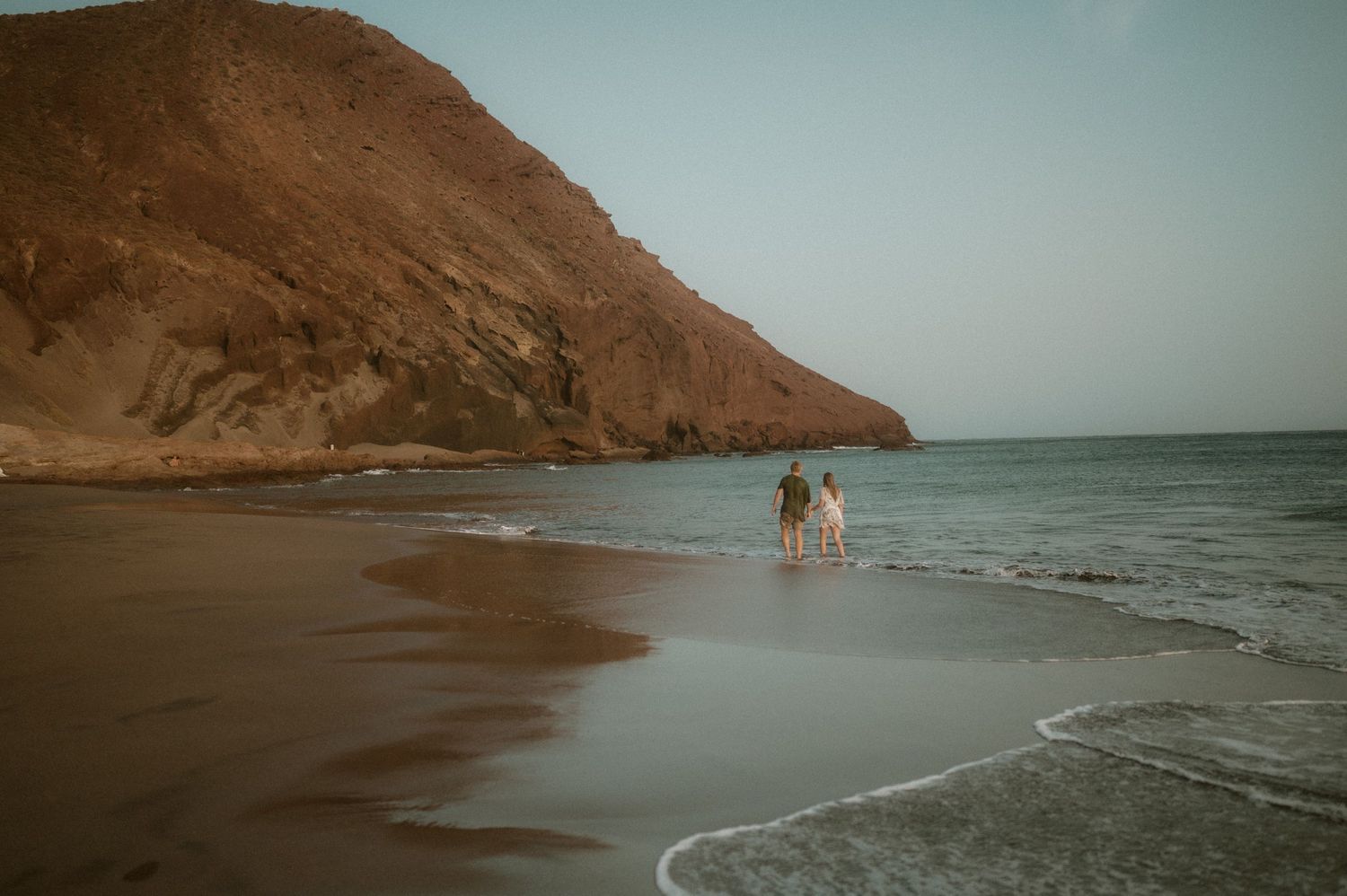 Couple standing hand in hand on Playa Tejita with ocean and Montaña Roja in the background