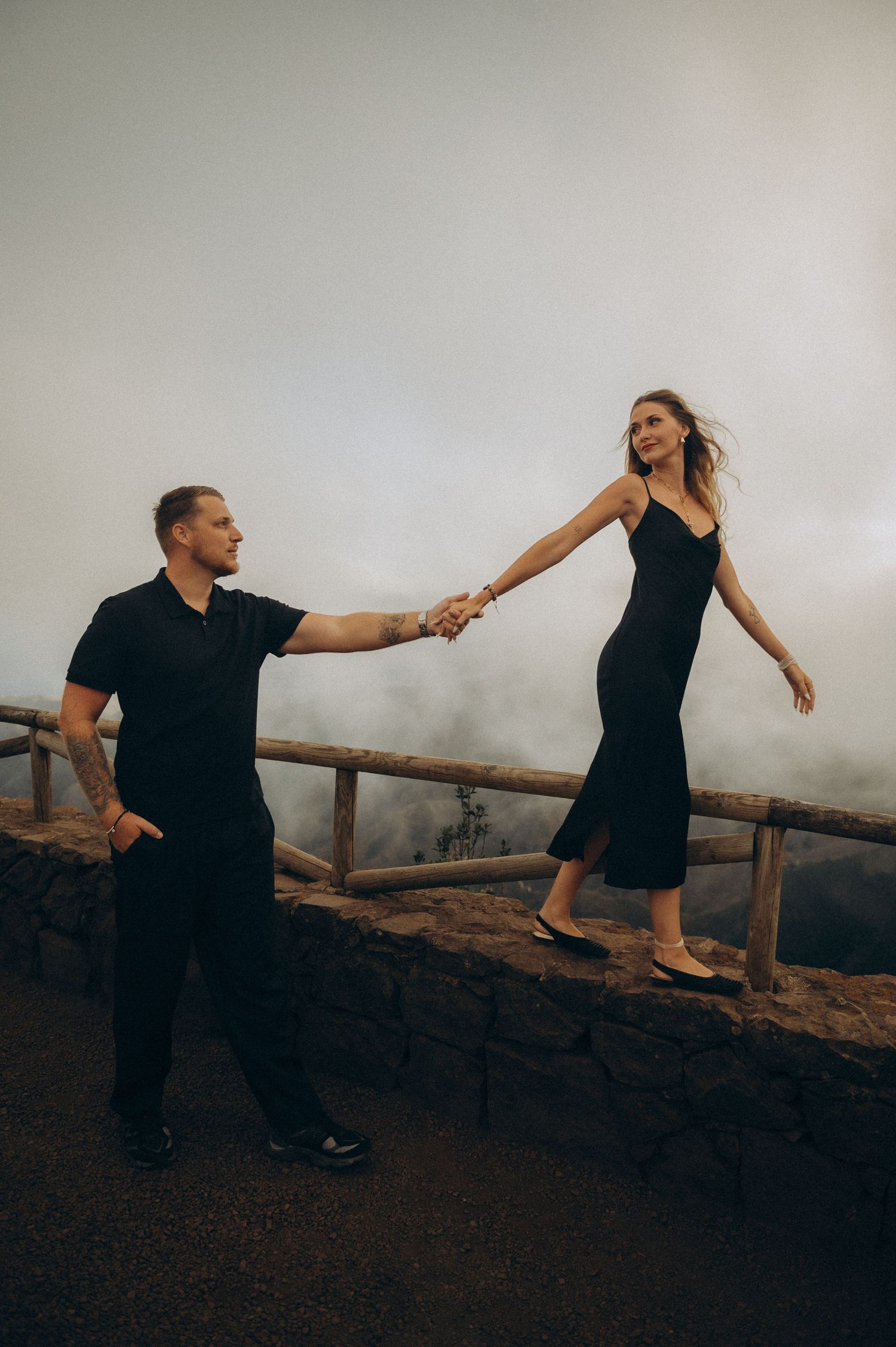 Couple in matching black outfits holding hands on a misty overlook with a rustic wooden fence.