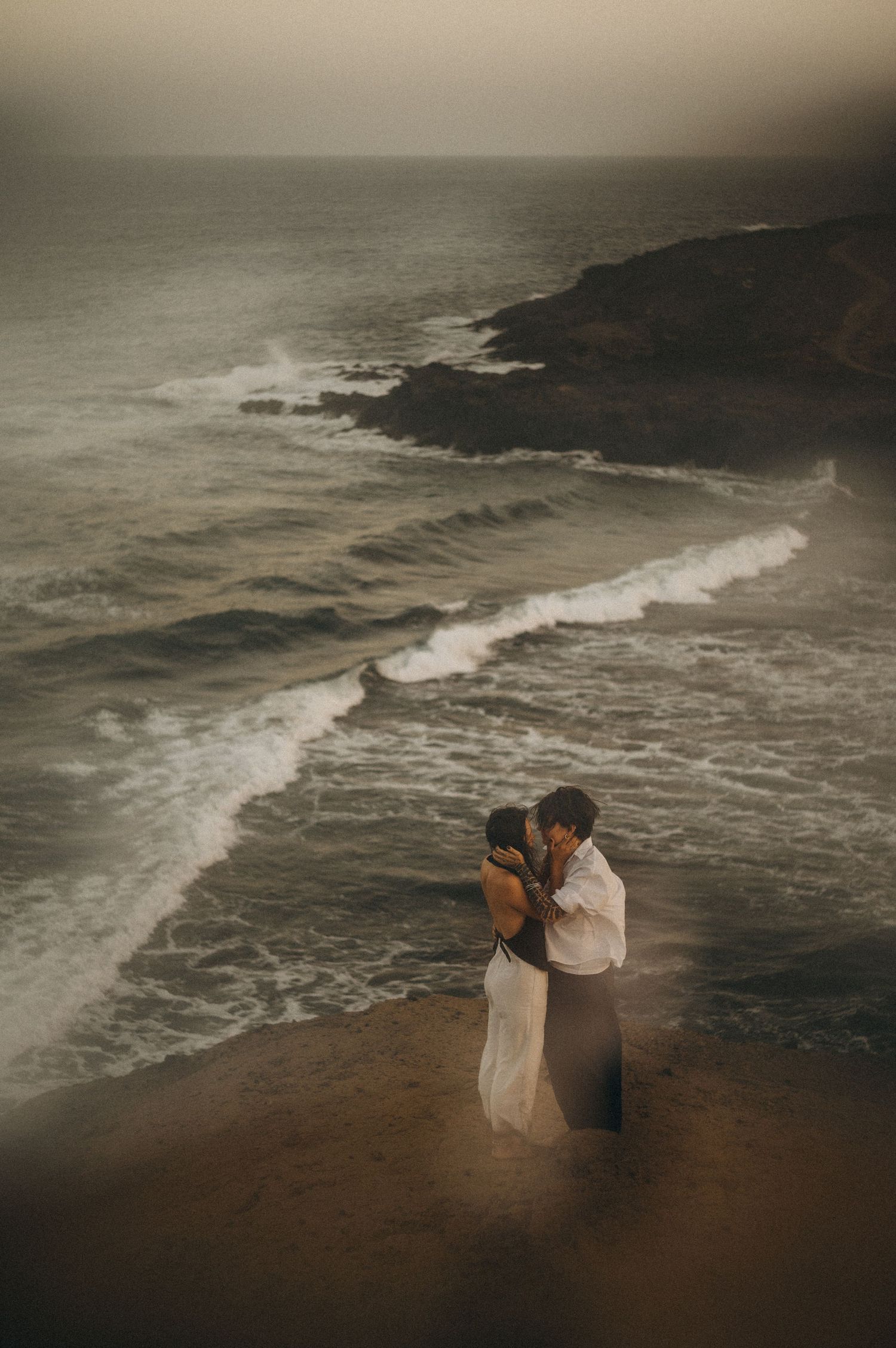 Couple embracing romantically on a misty coastal cliff overlooking dramatic ocean waves at dusk.