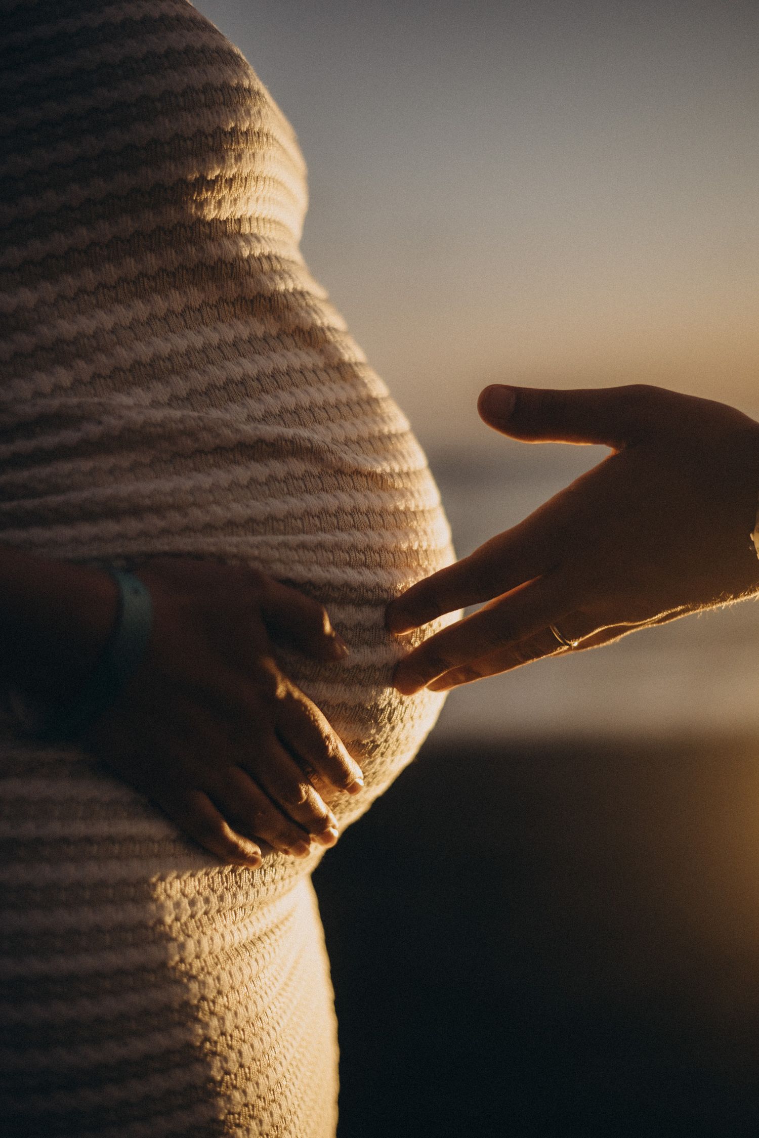 Pregnant woman in striped sweater touching baby bump at golden sunset, partner reaching out to touch belly.