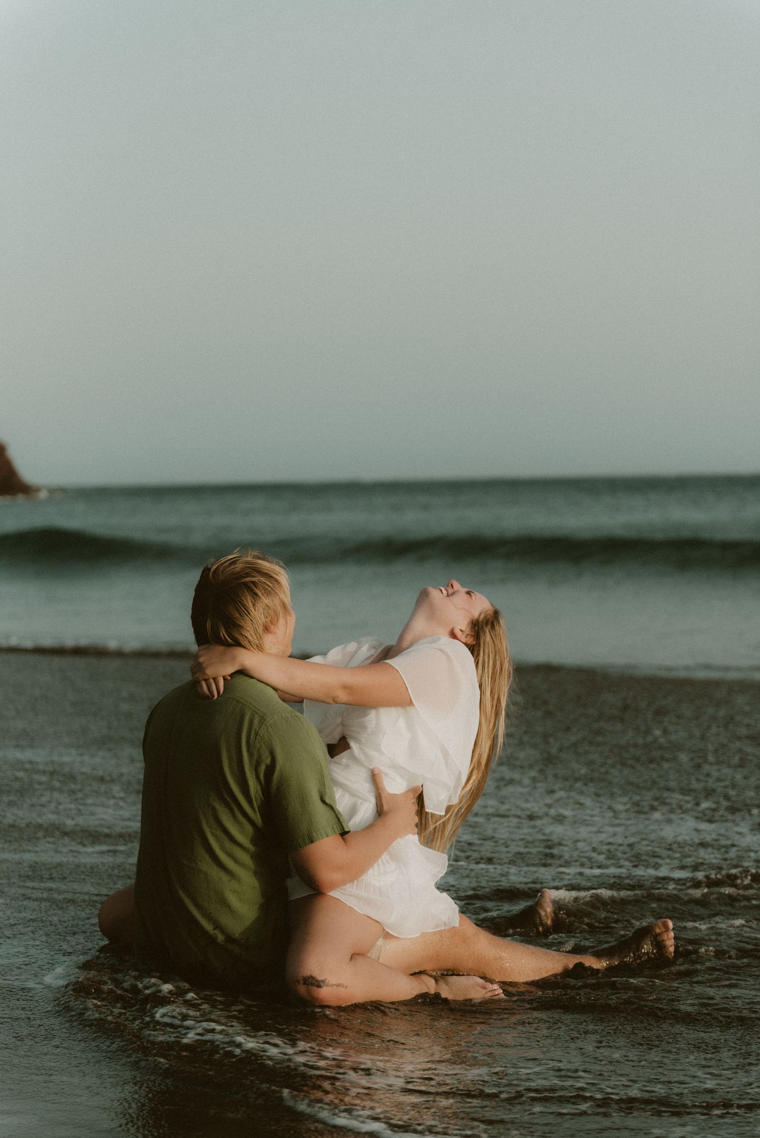 Young couple sitting embraced in the water on Playa Tejita, with ocean touching them, Tenerife couples photoshoot