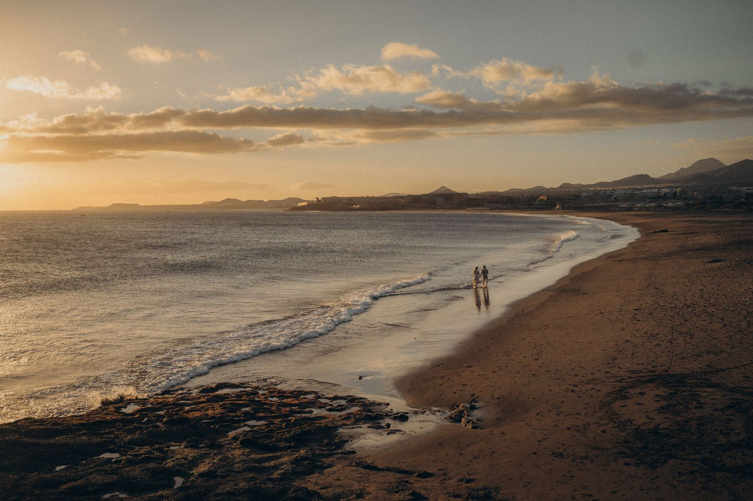 Dramatic coastal sunset panorama showing a curving beach shoreline with distant mountains and golden clouds over the ocean.