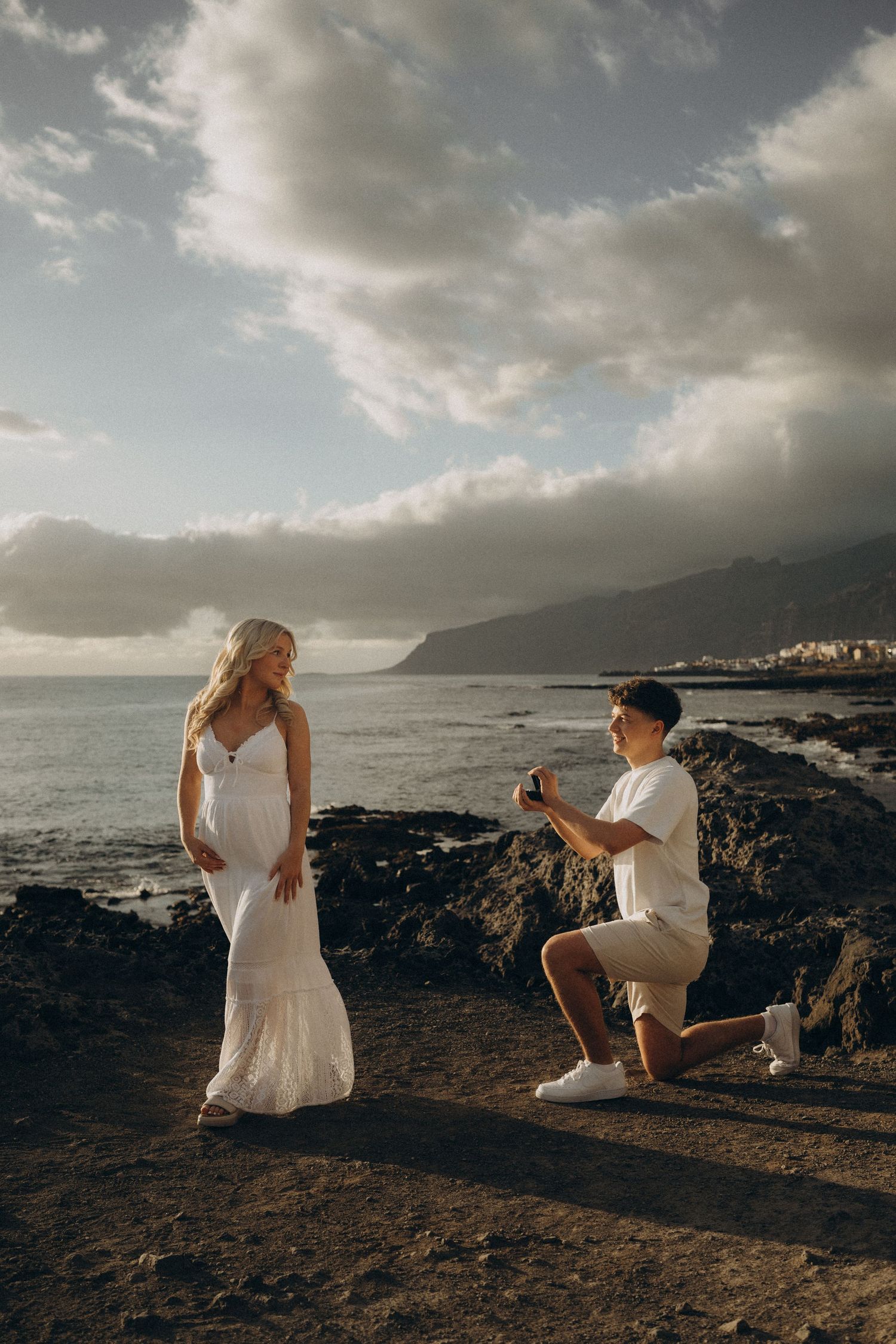 Man proposes to woman in white dress on rocky coastal cliff at sunset with ocean and mountains behind.