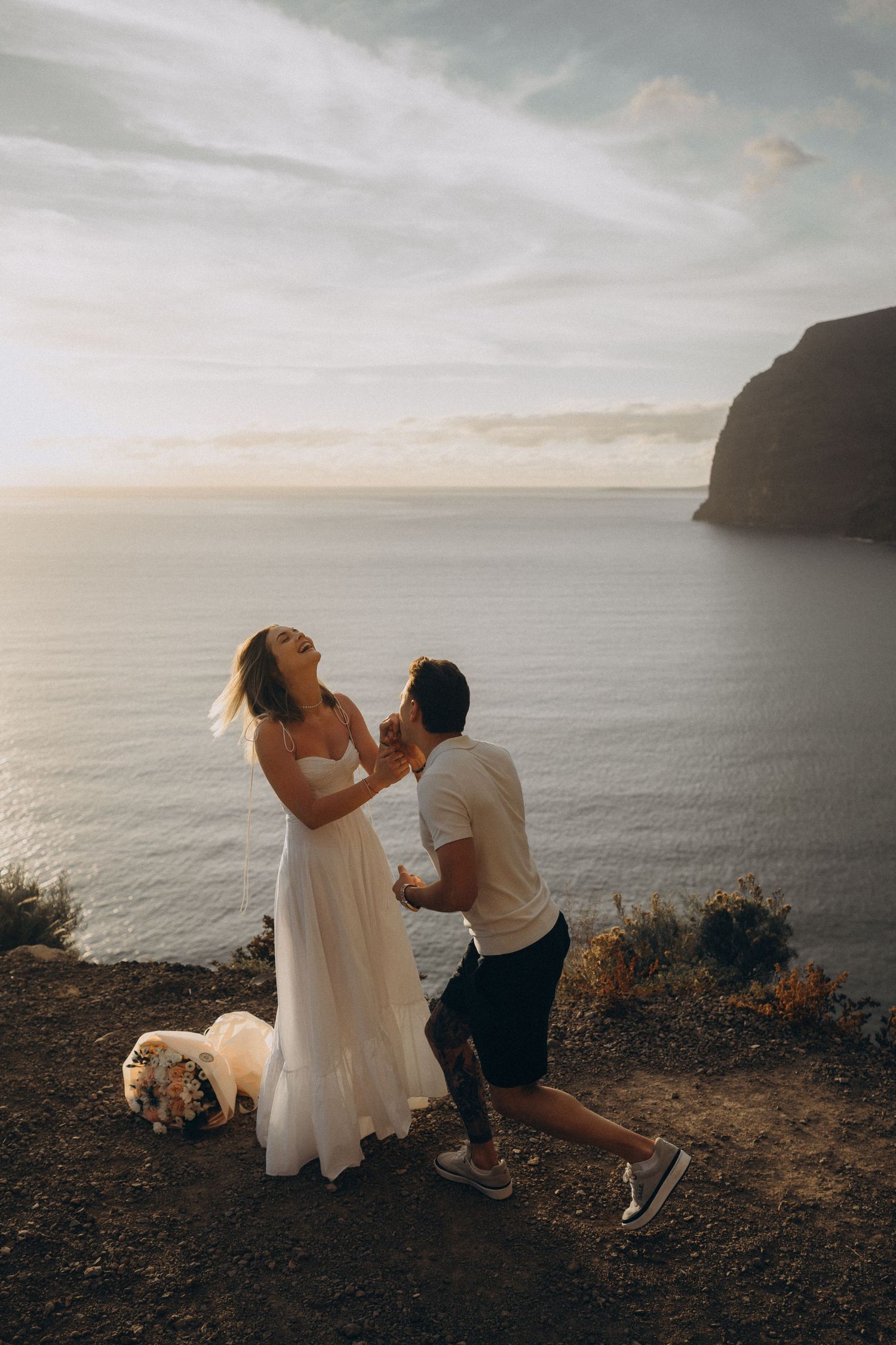 Man kneeling in proposal to woman in white dress on coastal cliff at sunset with ocean view.