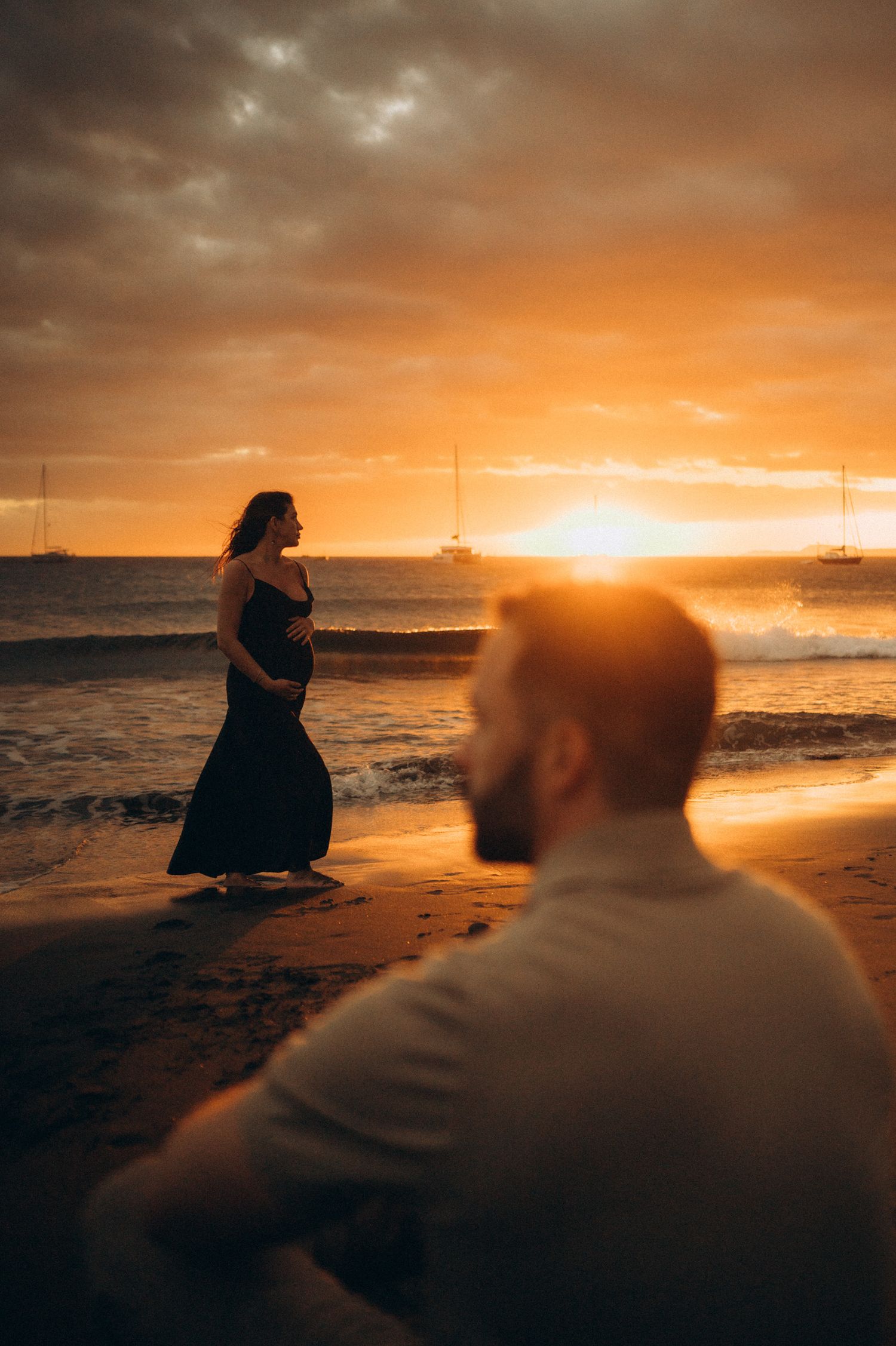 Pregnant woman in black dress stands on beach at golden sunset while man sits in foreground.